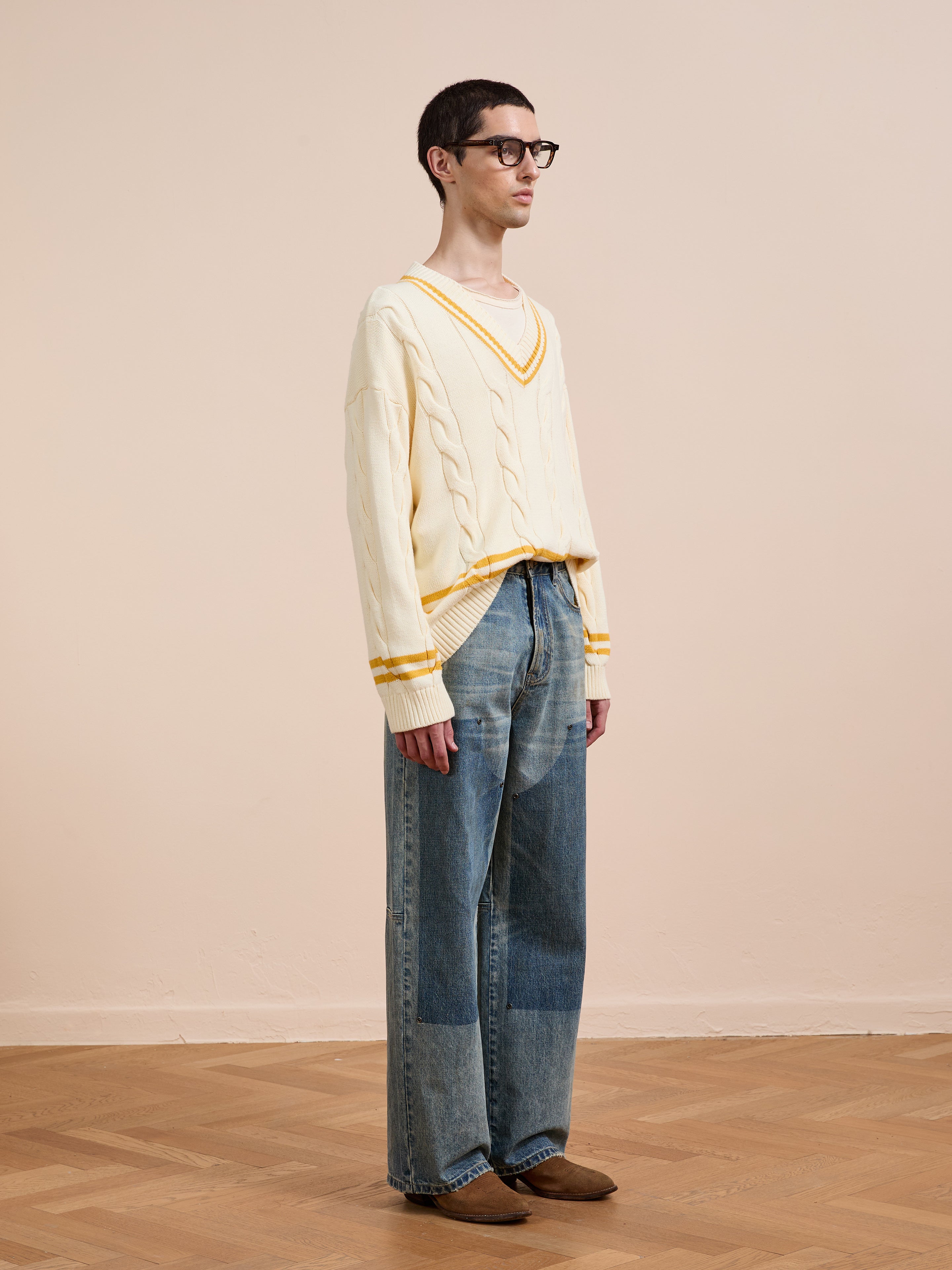 A person in glasses wears the FOUND V-Neck Cricket Sweater, featuring yellow trim, with loose patchwork jeans and brown shoes, standing on a wooden floor in a neutral indoor space.