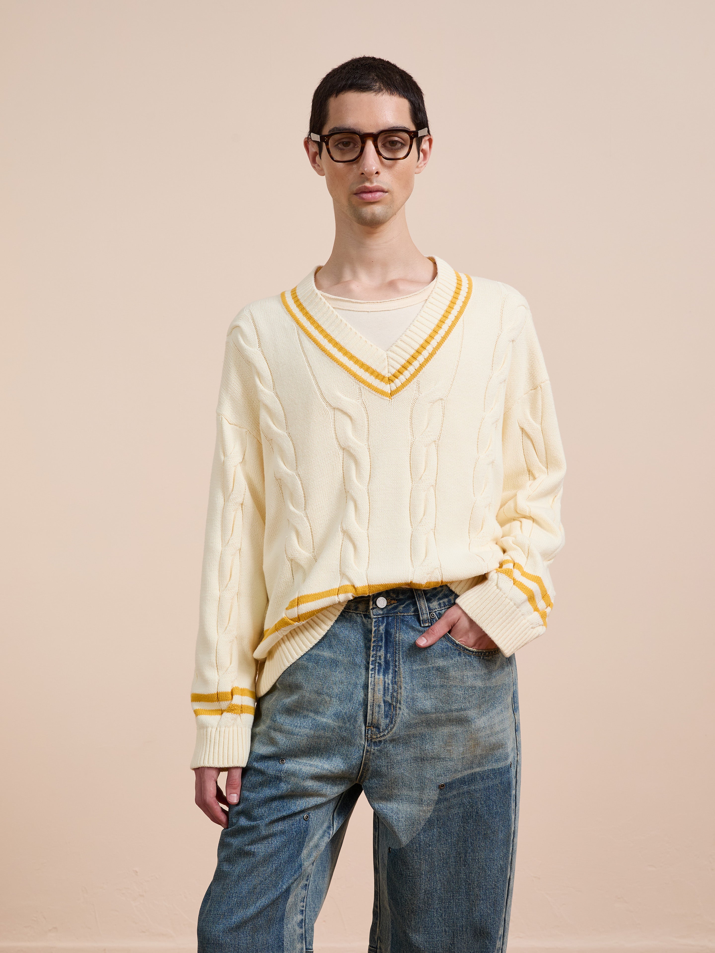 A person in FOUND's V-Neck Cricket Sweater—a cream unisex knit with yellow stripes—paired with blue jeans and glasses stands against a plain beige background.