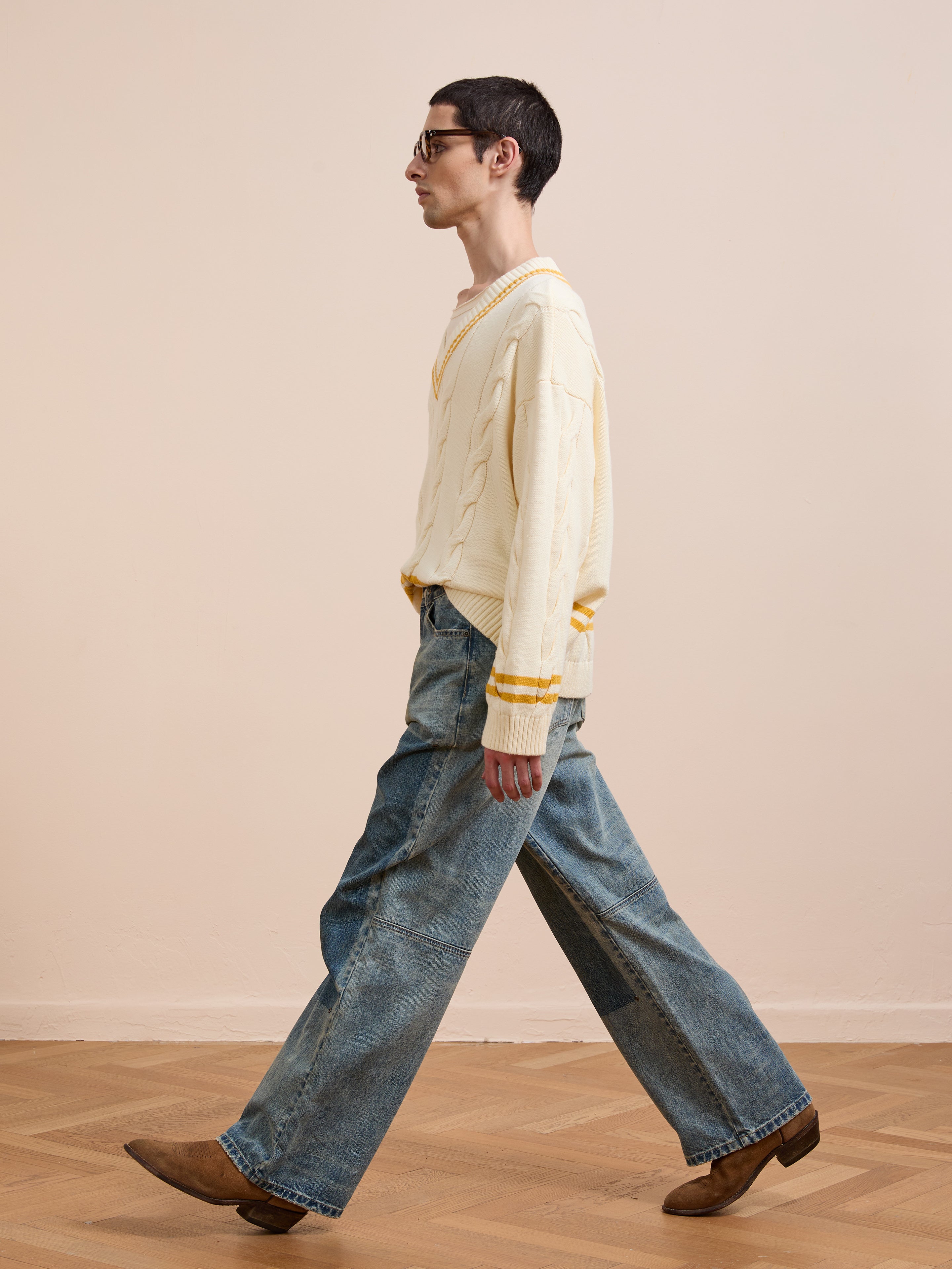 A person in the FOUND V-Neck Cricket Sweater, wide-leg blue jeans, and brown shoes walks indoors against a plain beige wall.