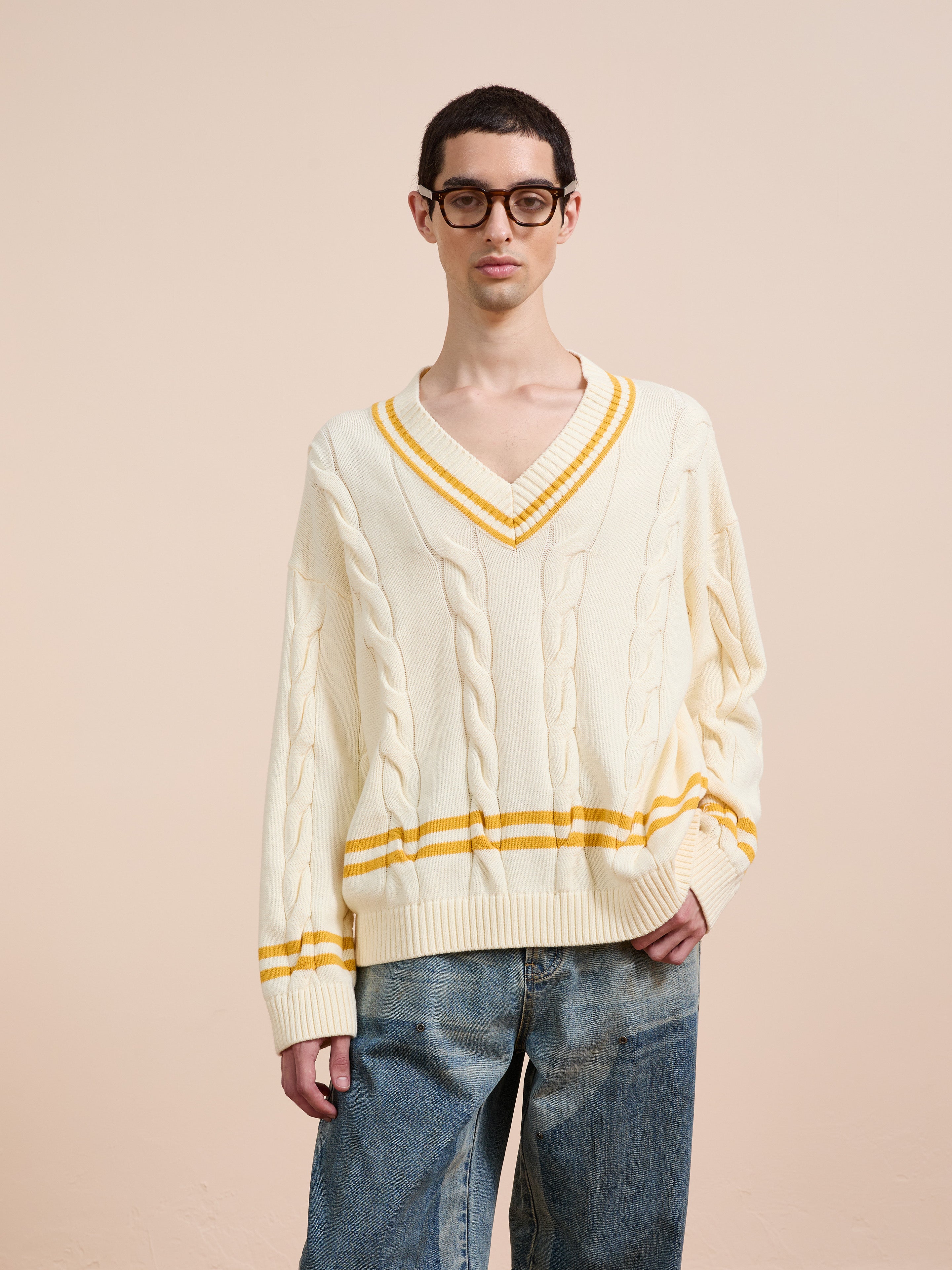 A person wearing glasses and the FOUND V-Neck Cricket Sweater with yellow stripes, paired with loose blue jeans, stands against a plain beige background.