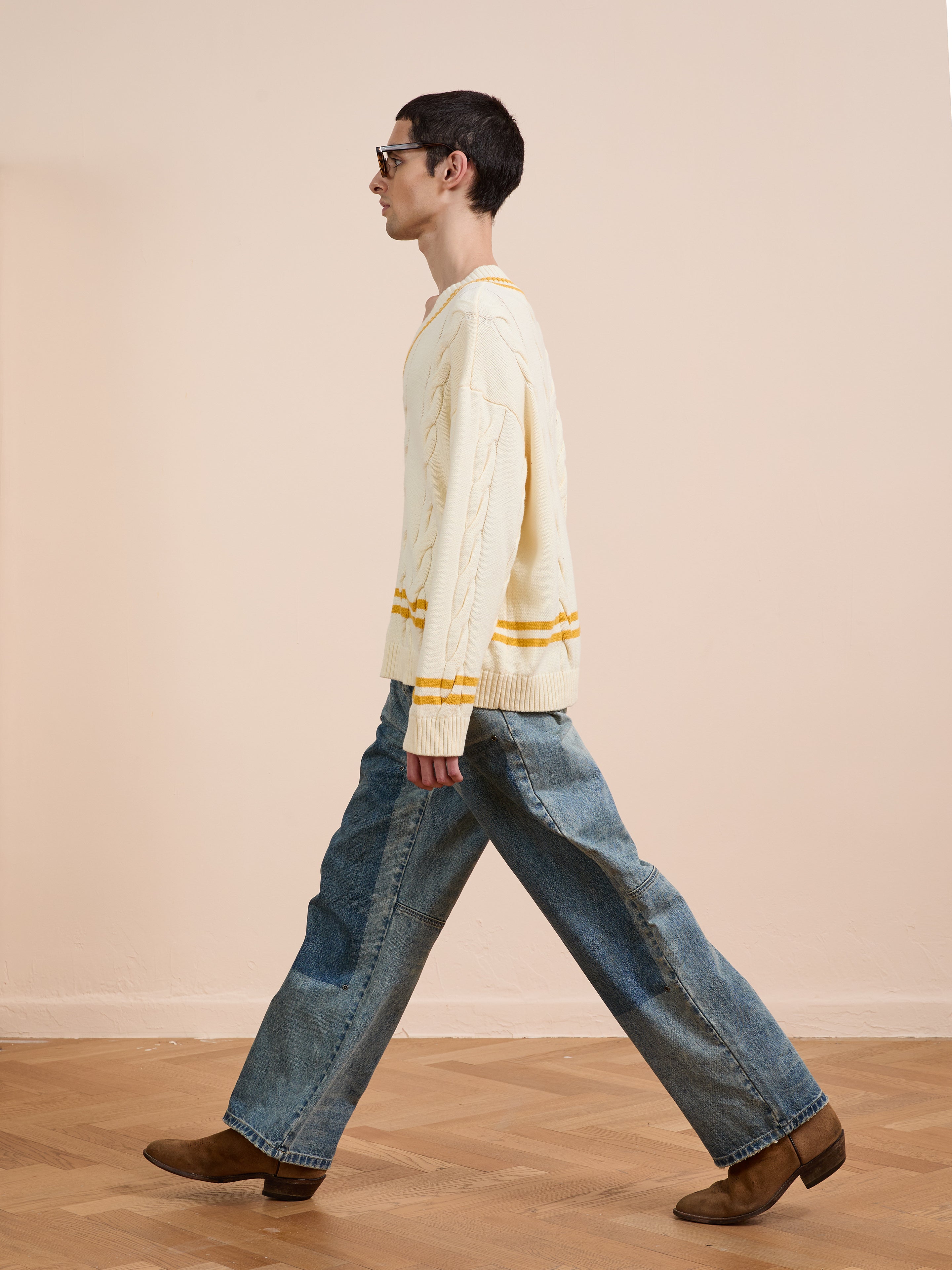 A man wears the FOUND V-Neck Cricket Sweater, featuring yellow stripes, paired with blue wide-leg jeans and brown boots as he walks on a wooden floor against a plain beige background.