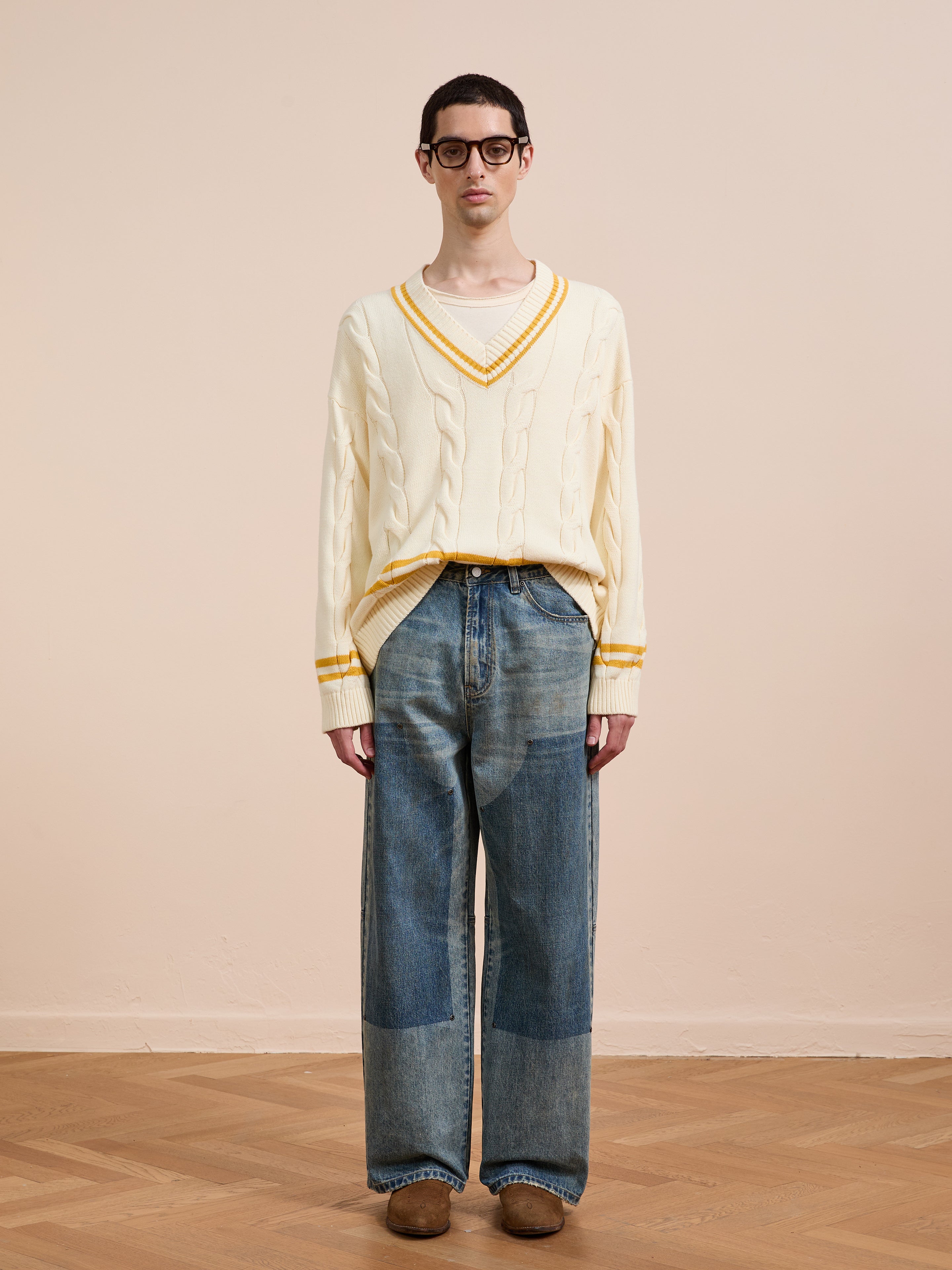 A person wearing glasses, a FOUND V-Neck Cricket Sweater with yellow details, loose-fitting patchwork jeans, and brown shoes stands on a wooden floor against a plain beige background.