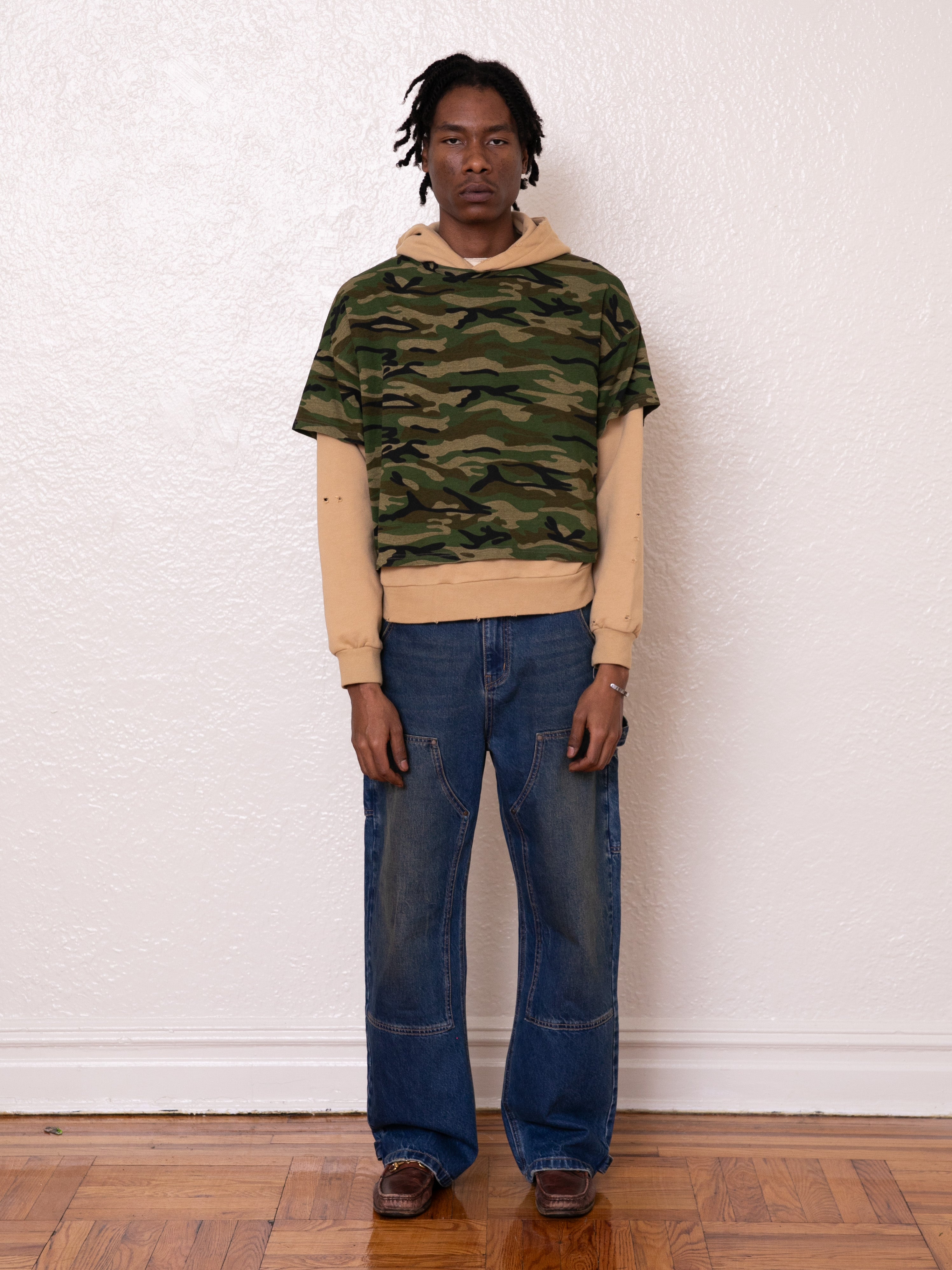 A person stands against a white wall wearing a camouflage shirt over the Double Layer Hoodie by FOUND in beige, paired with vintage-look blue jeans and brown shoes.