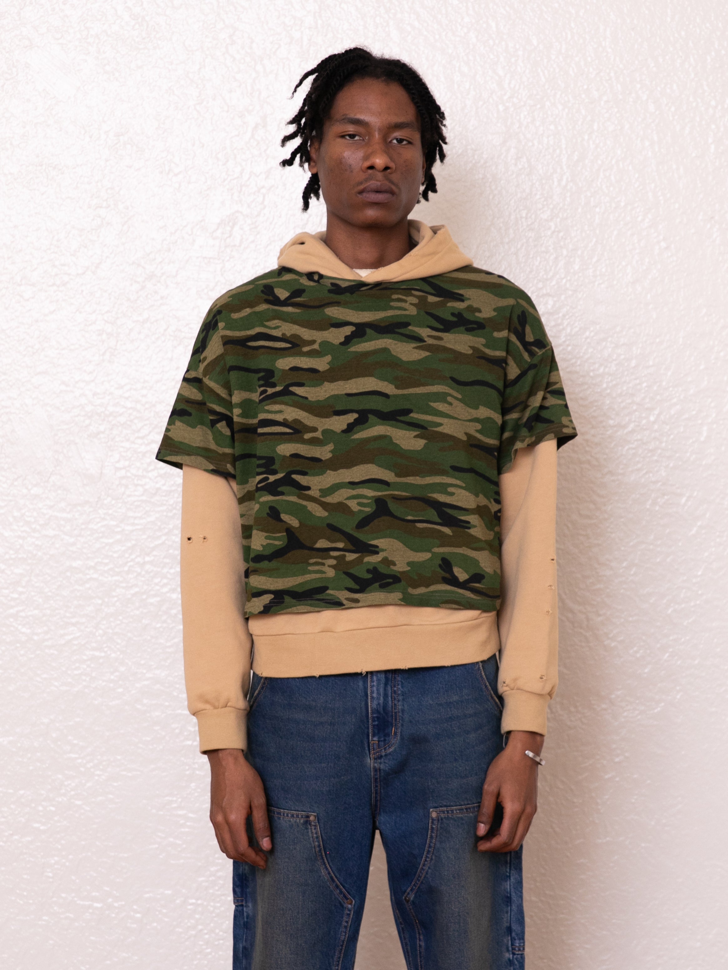 A person is wearing a camouflage short-sleeve over the Double Layer Hoodie by FOUND, which is crafted from French terry cotton, paired with blue jeans. They stand against a white textured wall, effortlessly pulling off a vintage look.