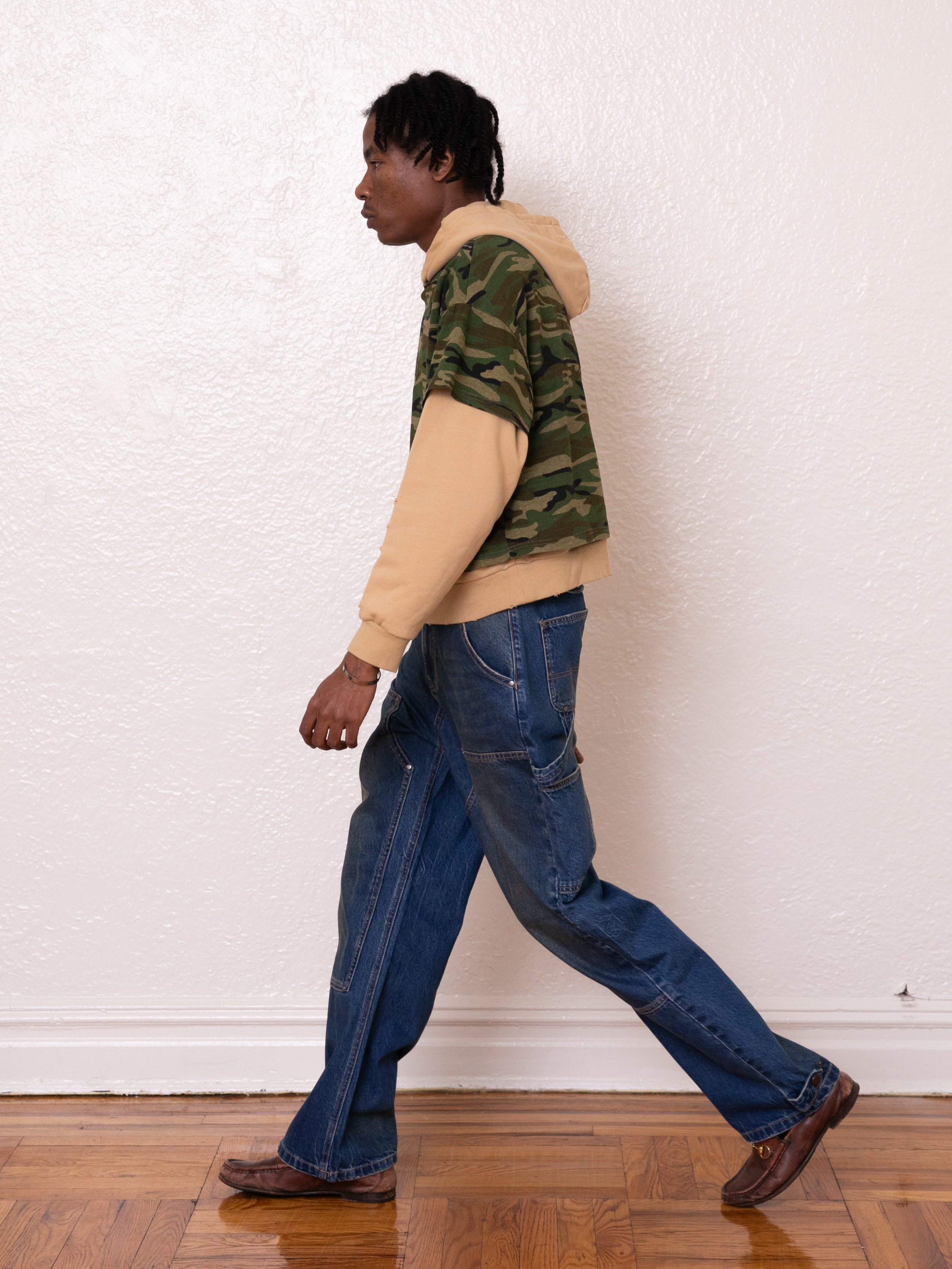 A person strolls indoors wearing a FOUND Double Layer Hoodie made from soft French terry cotton, paired with jeans and brown shoes. The camouflage pattern of the outfit subtly contrasts against the plain white wall, adding a touch of vintage appeal.