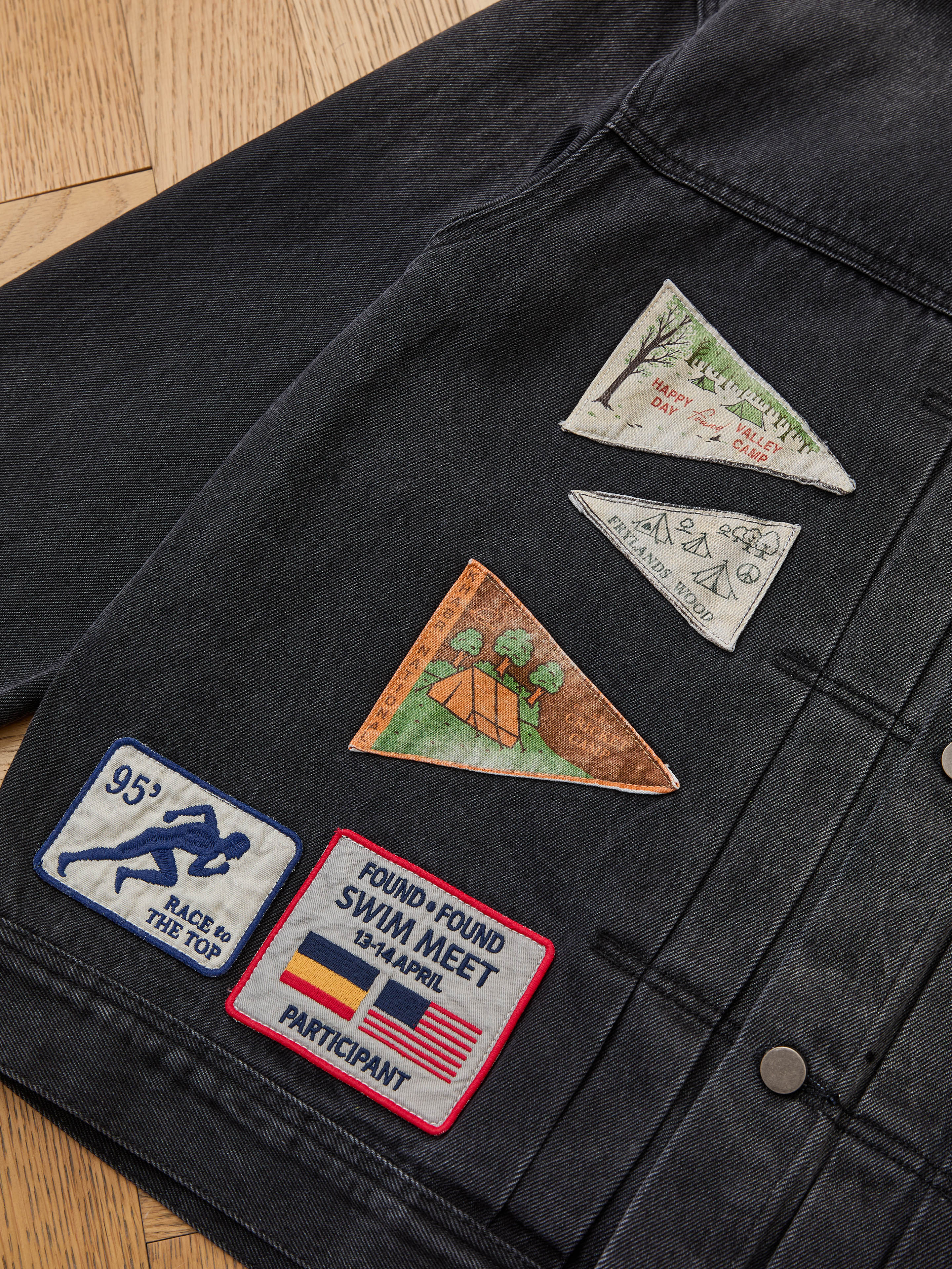The FOUND Denim Patchwork Jacket boasts a corduroy collar and five eye-catching Americana embroidered patches, including camping scenes and badges, all showcased on a wooden floor.