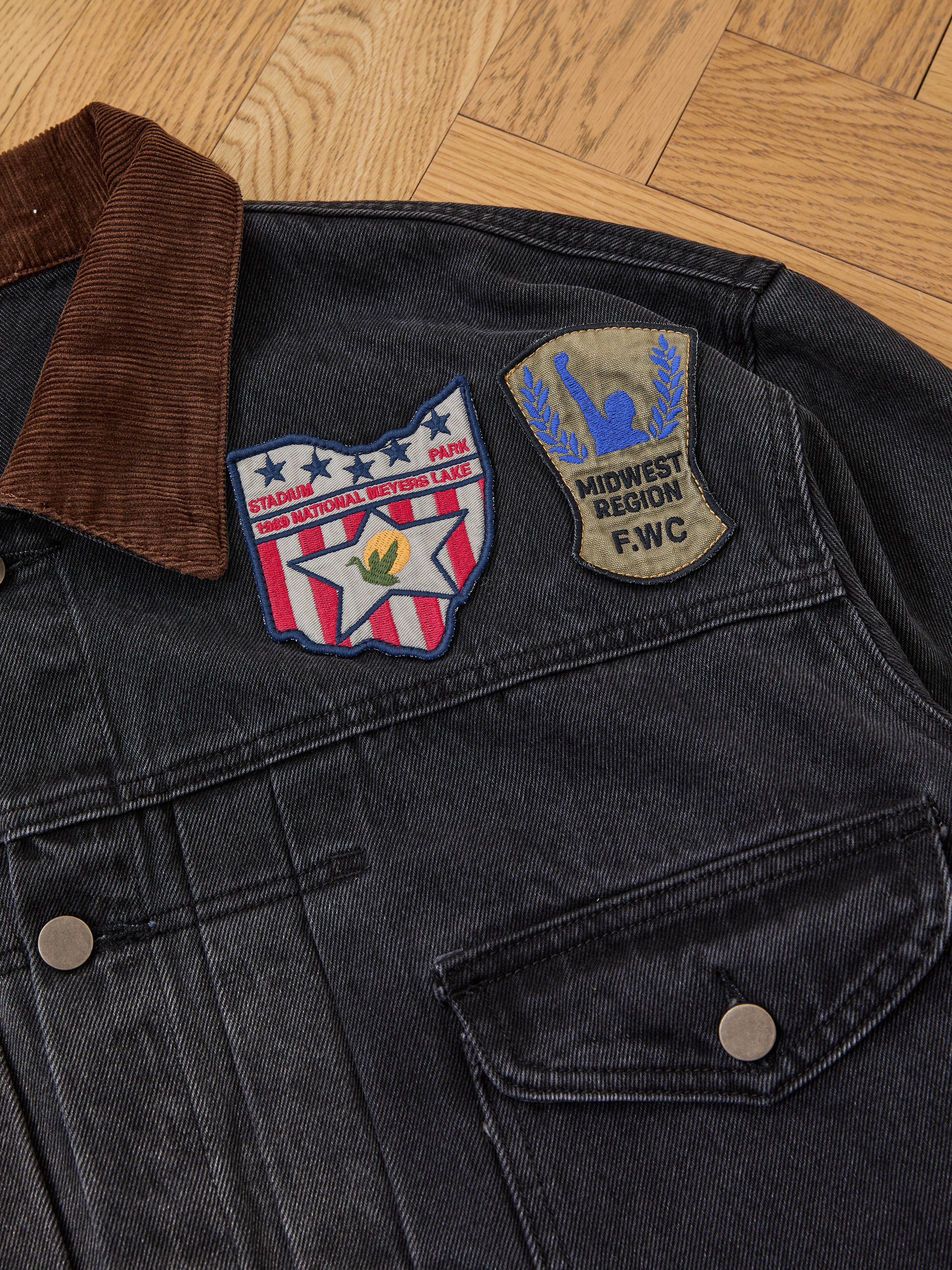 The FOUND Denim Patchwork Jacket features metal buttons, a corduroy collar, and two Americana patches: a red, white, and blue shield and a khaki "Midwest Region FWC" patch.