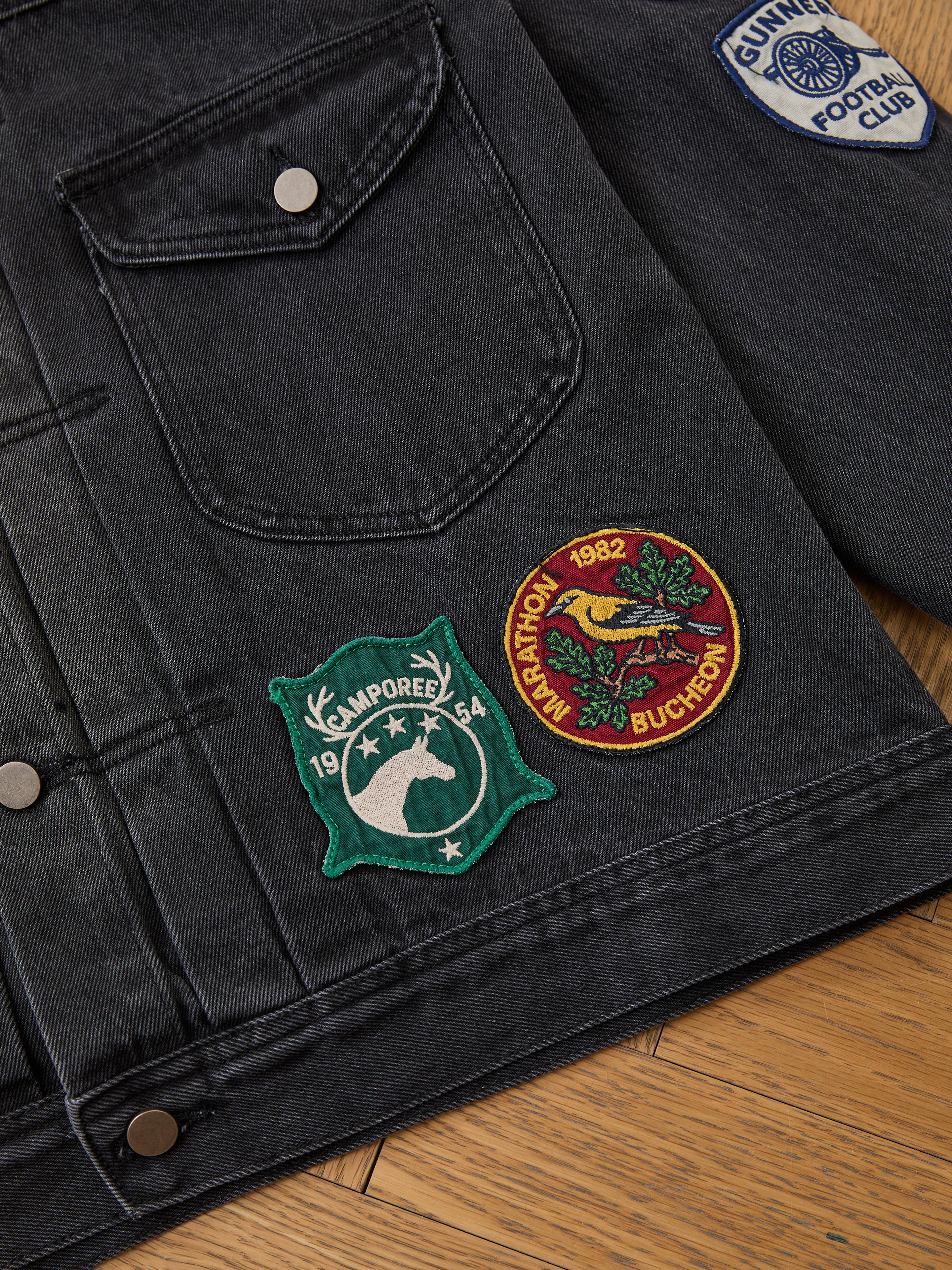 The FOUND Denim Patchwork Jacket features a corduroy collar and three Americana patches—a green badge, red marathon patch, and blue football club badge—displayed on a wooden floor.