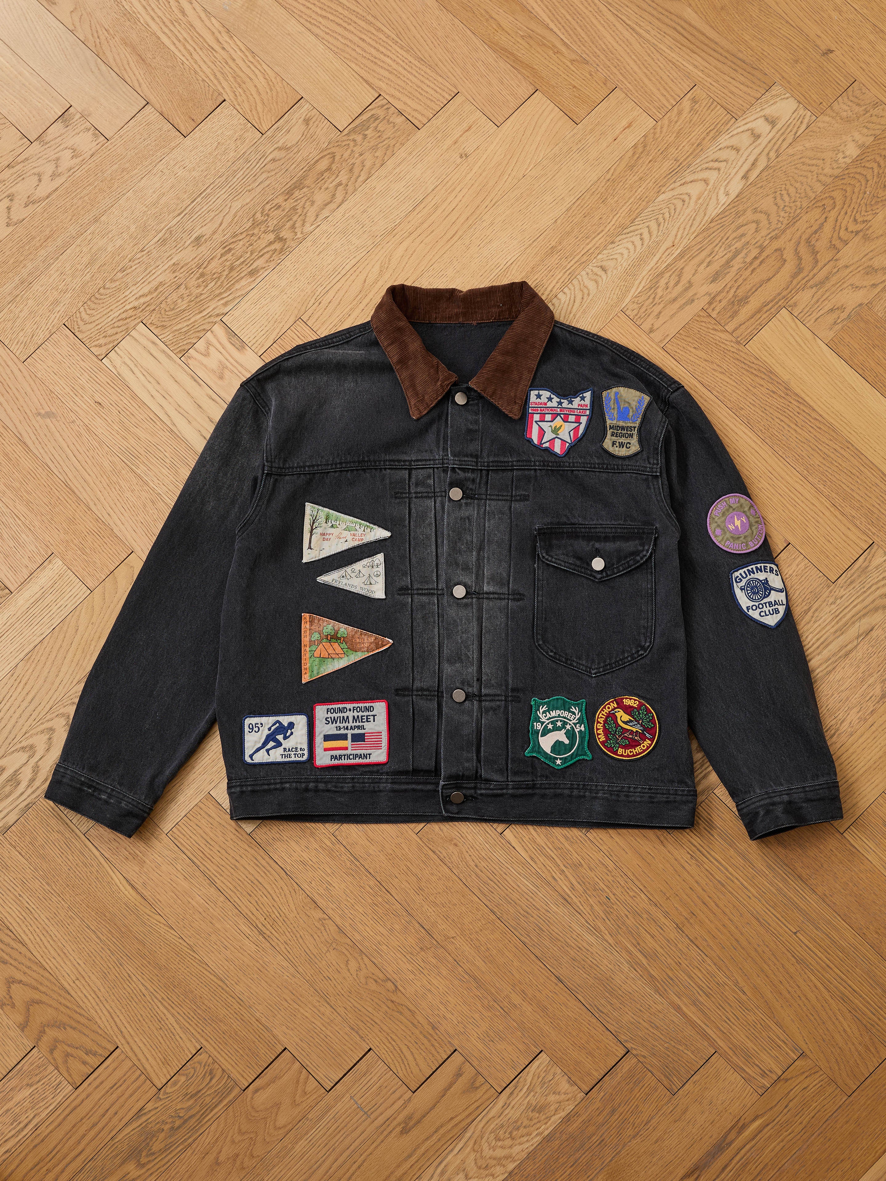 The FOUND Denim Patchwork Jacket features a black vintage wash, corduroy collar, and colorful Americana patches and badges on the front and right sleeve, displayed flat on a wooden herringbone floor.