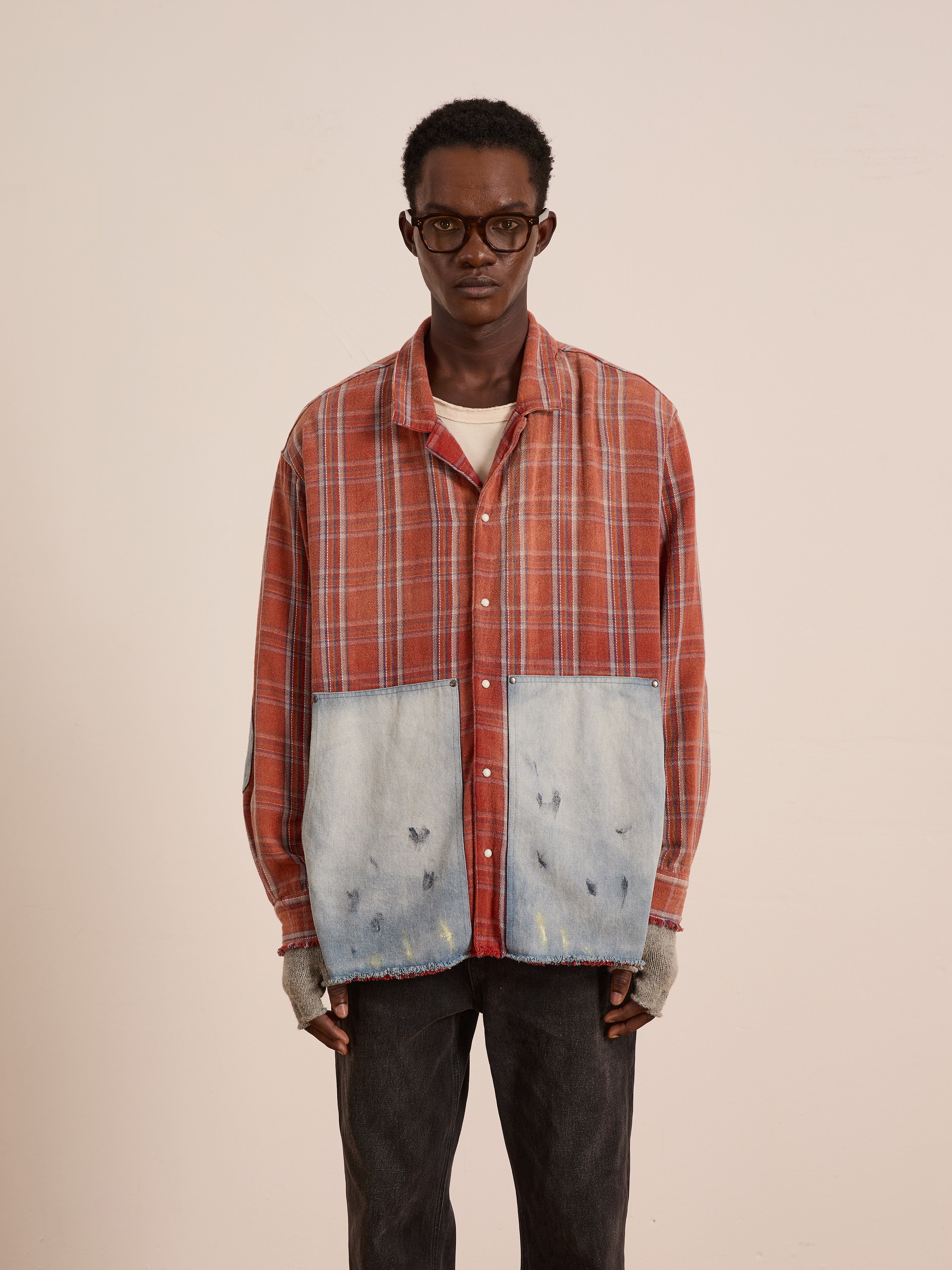 A man in glasses wears the FOUND Plaid Denim Shirt—an orange unisex relaxed-fit shirt with large faded denim carpenter pockets—over a white tee, paired with dark pants and gray fingerless gloves, standing against a plain light background.
