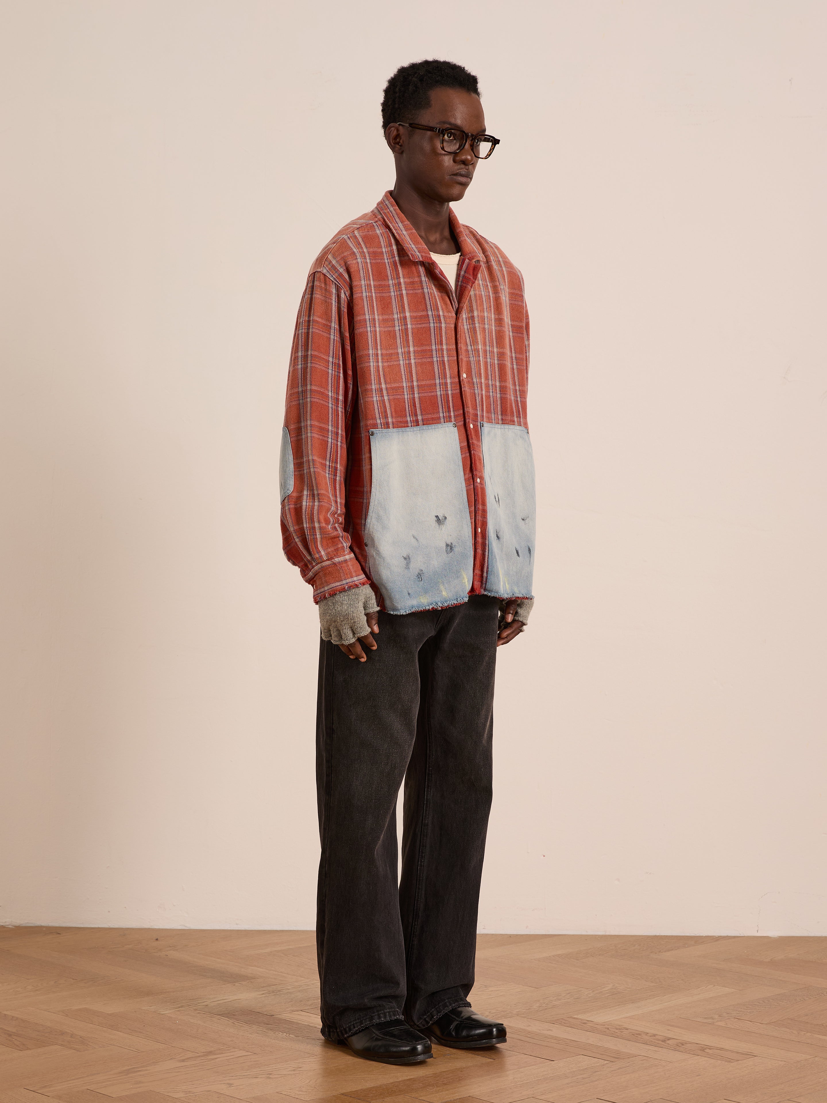 A person wears the FOUND Plaid Denim Shirt—a unisex, relaxed-fit red plaid top with light blue denim carpenter pockets—paired with dark pants, black shoes, and glasses, standing against a plain light-colored wall.