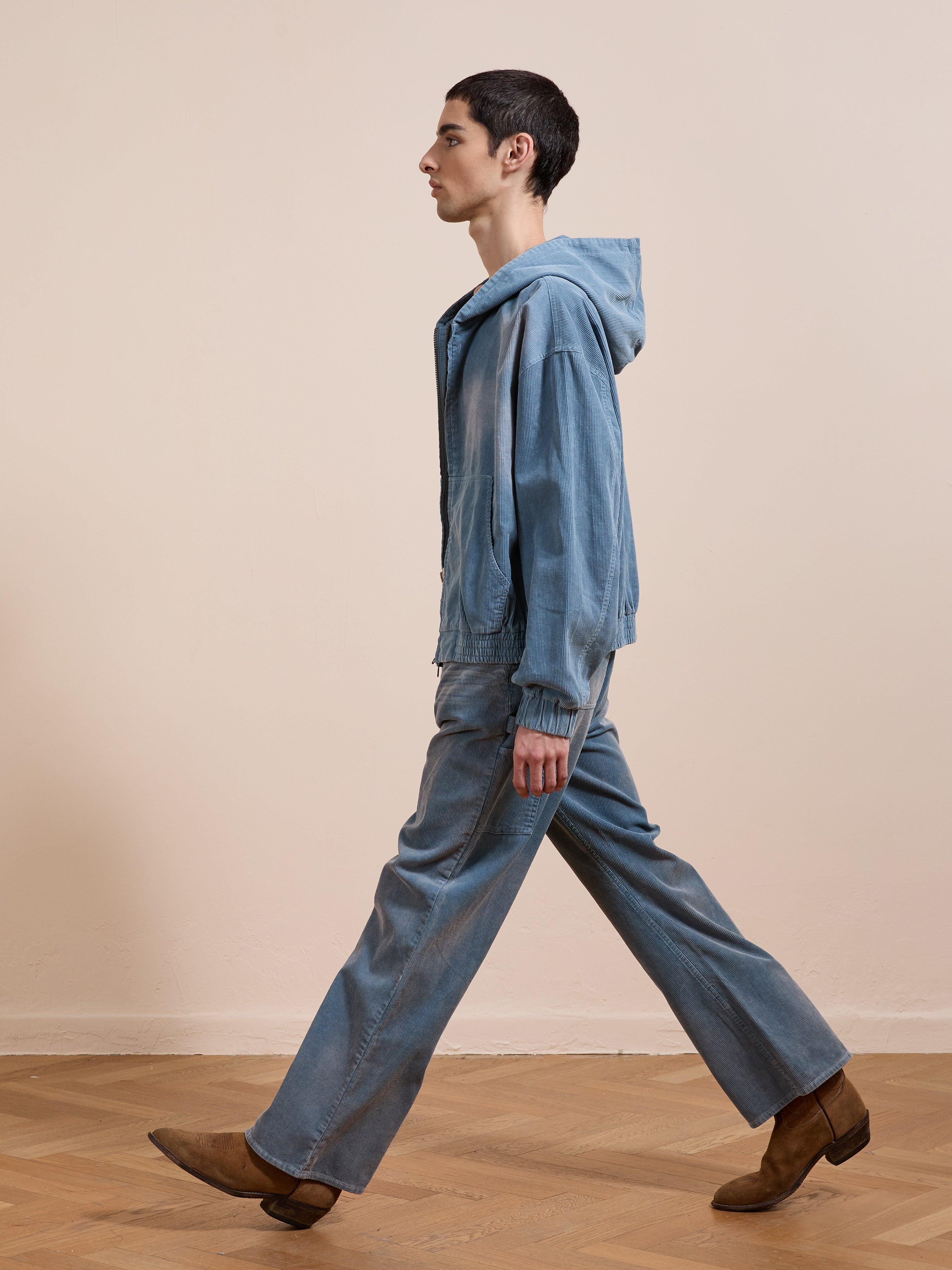 A person wearing the FOUND Corduroy Zip Up Hoodie and jeans, paired with brown boots, walks indoors on a wooden floor against a plain beige wall.