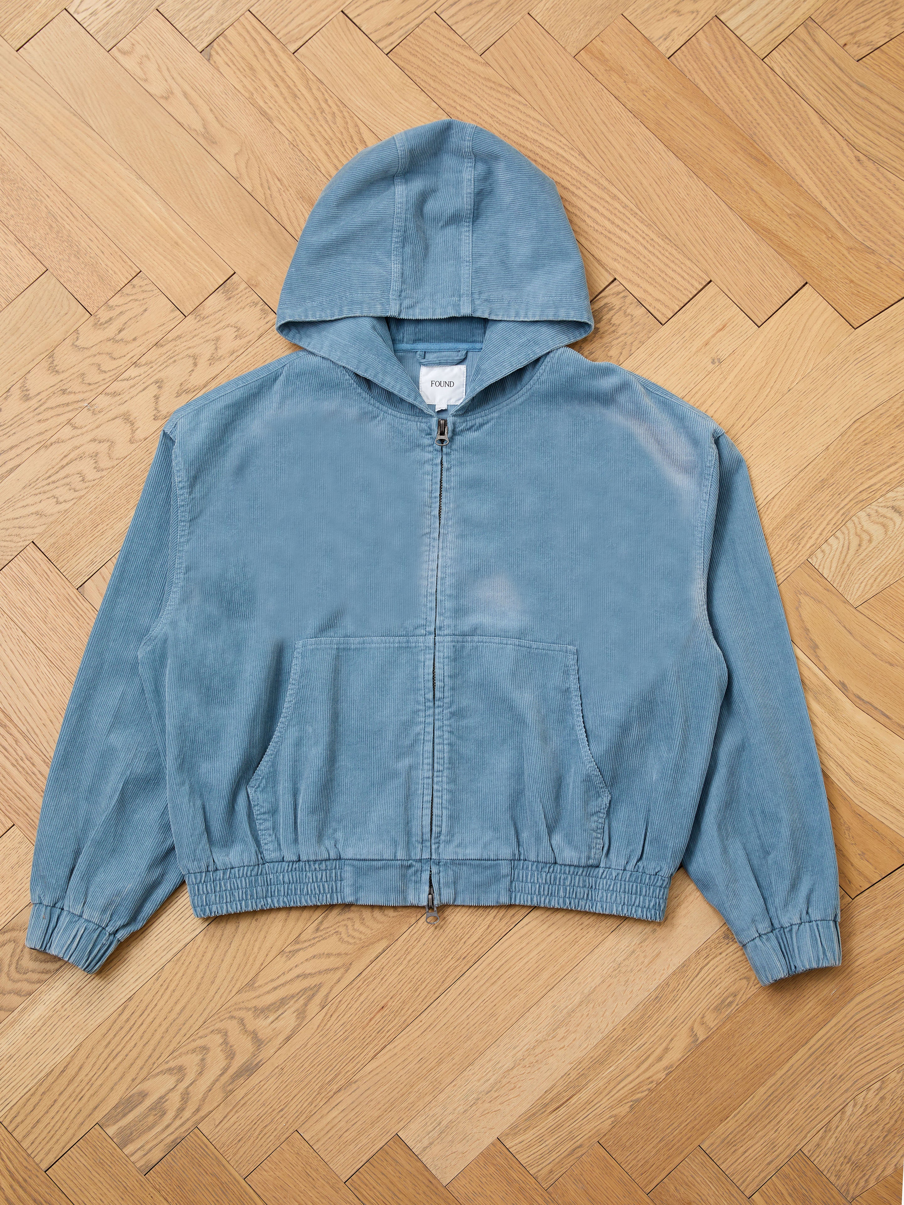 The FOUND Corduroy Zip Up Hoodie in faded blue features an elastic waistband and cuffs, front pockets, and a plain label, shown laid flat on a herringbone wood floor.