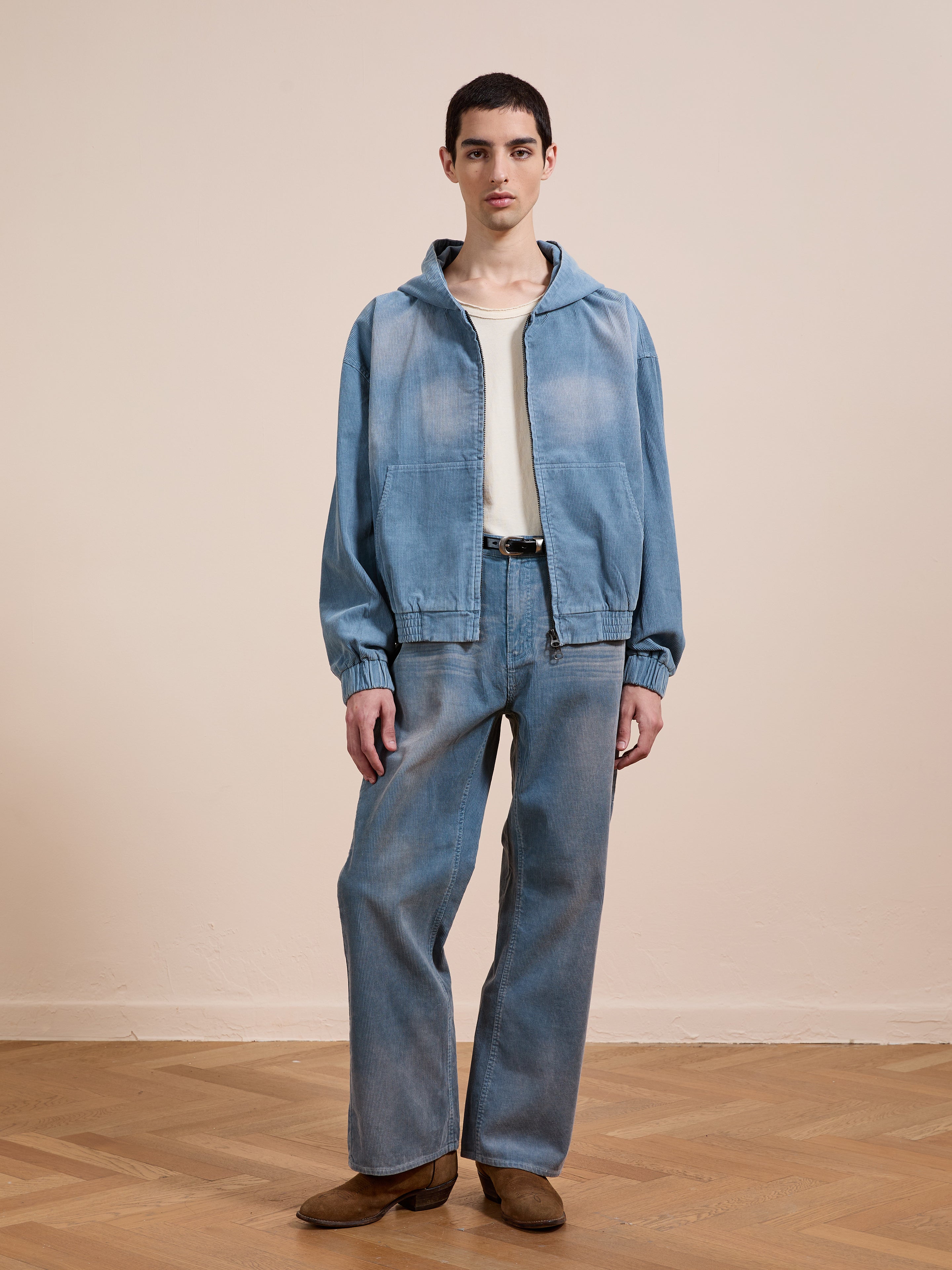 Indoors, a person wears a light-wash denim jacket and jeans, white shirt, black belt, brown shoes, and the FOUND Corduroy Zip Up Hoodie, standing against a beige wall with a wooden floor.