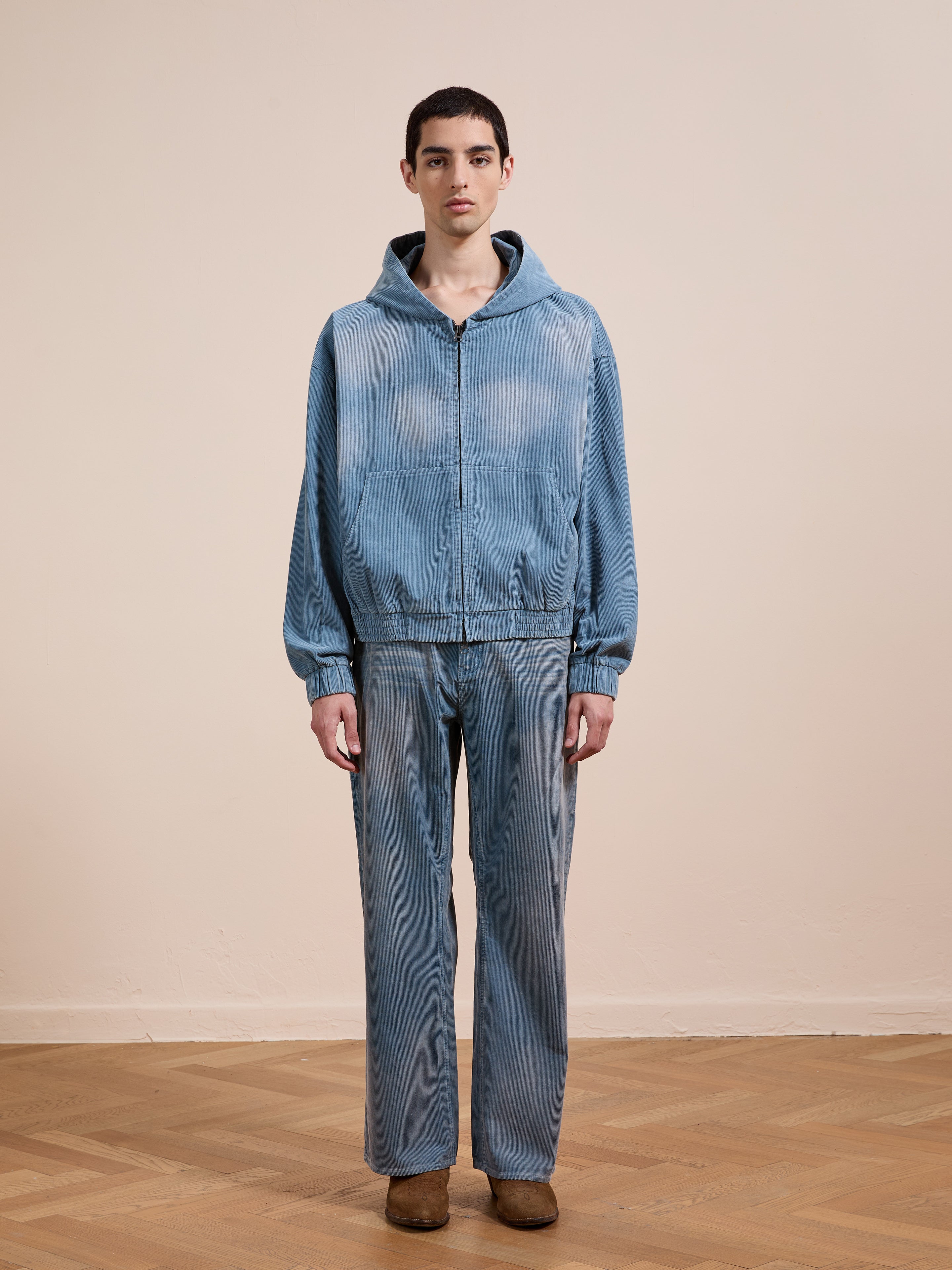 Indoors, a person wears the FOUND Corduroy Zip Up Hoodie with matching jeans and brown shoes, standing against a plain beige wall and wooden floor.