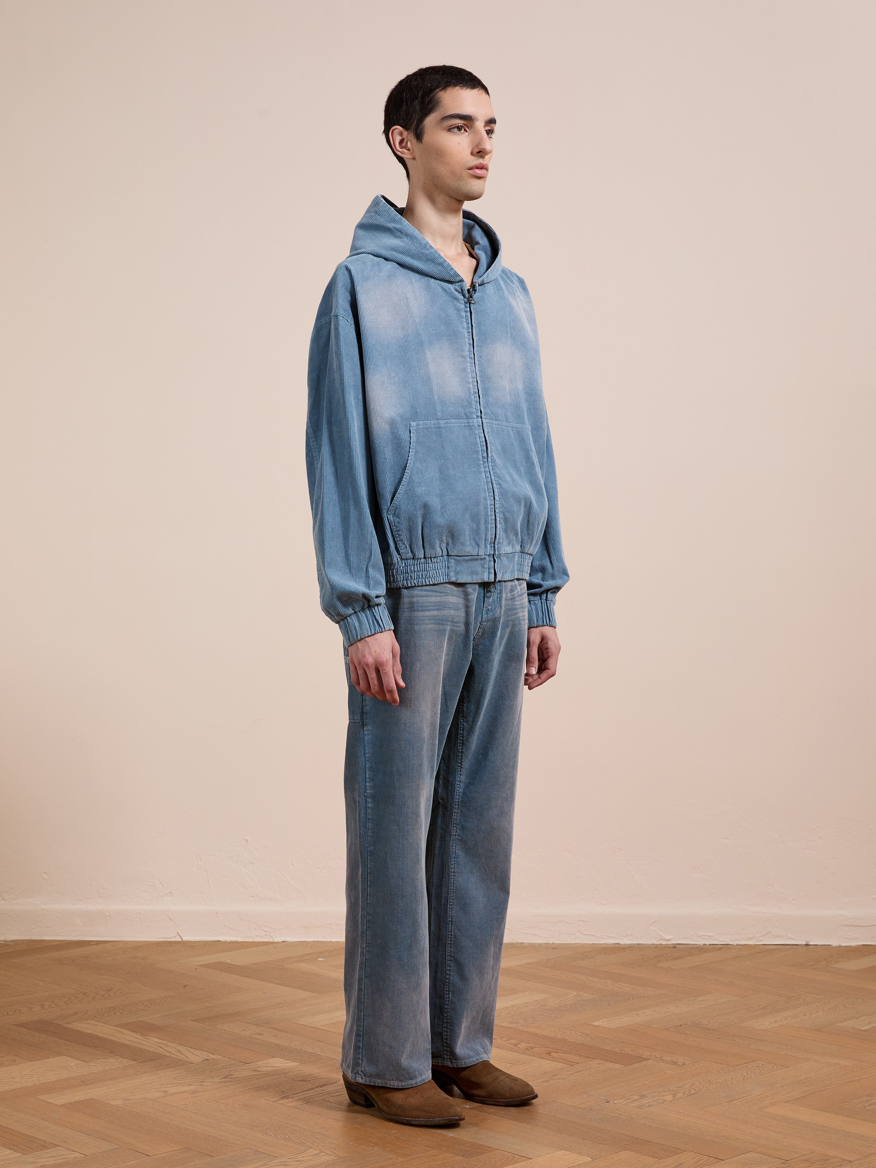 A person stands indoors on a wooden floor wearing the FOUND Corduroy Zip Up Hoodie in faded blue, matching denim pants, and brown shoes, facing slightly to the right.