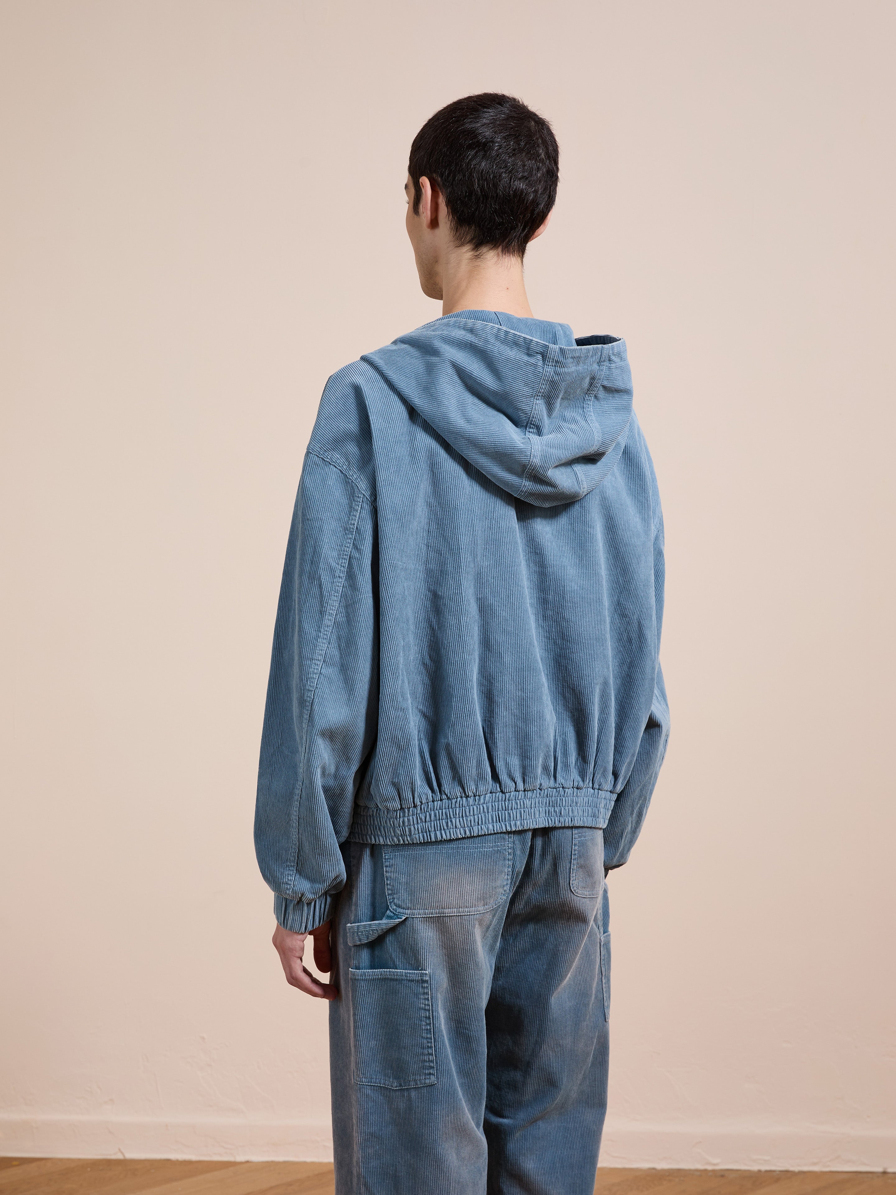 A person stands indoors facing away from the camera, wearing a FOUND Corduroy Zip Up Hoodie in faded blue with matching blue pants featuring a pocket.