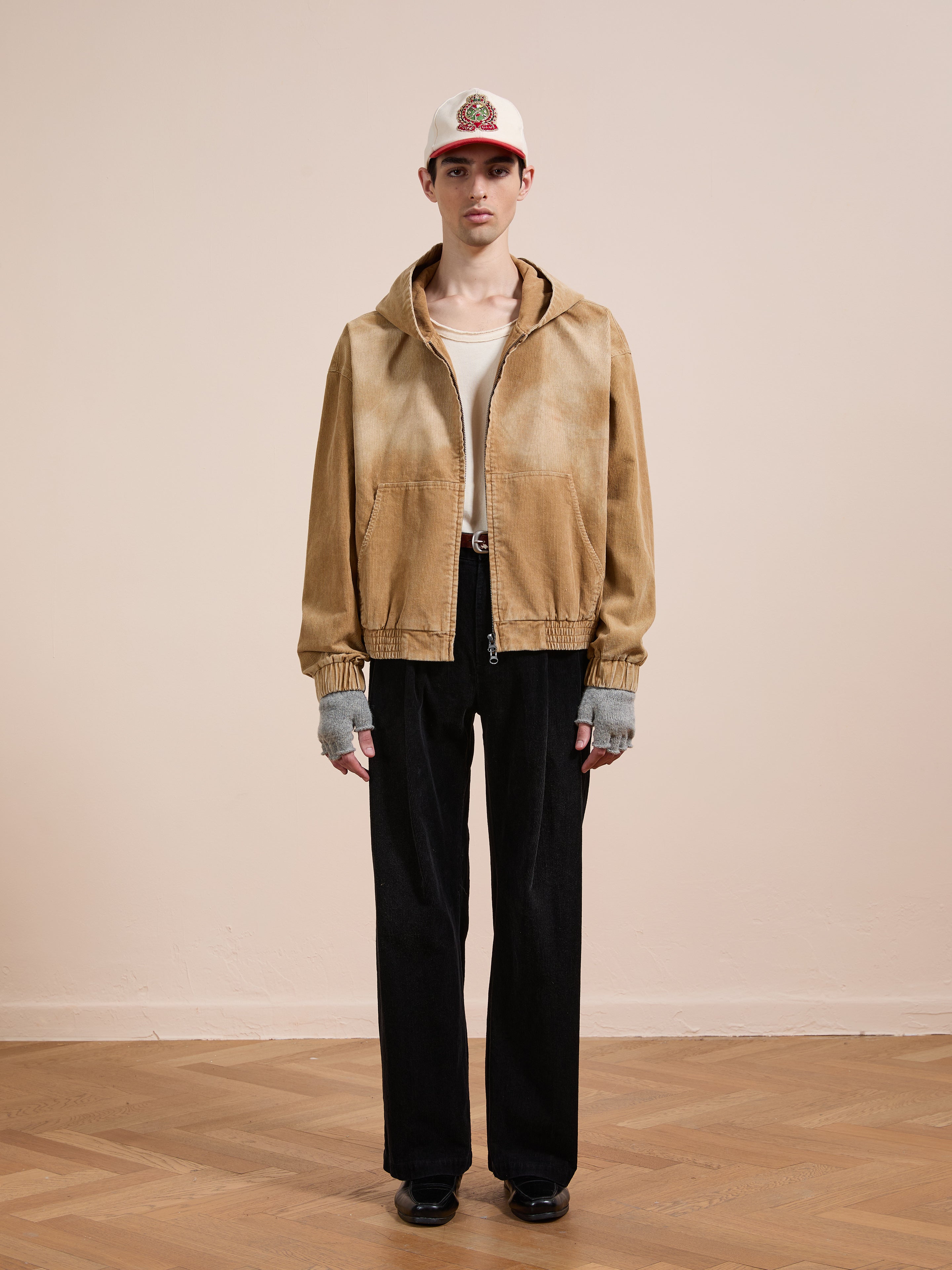 A person stands indoors wearing the FOUND Corduroy Zip Up Hoodie with a vintage vibe, paired with black pants, gray fingerless gloves, a white tee, and a white baseball cap with a red brim.