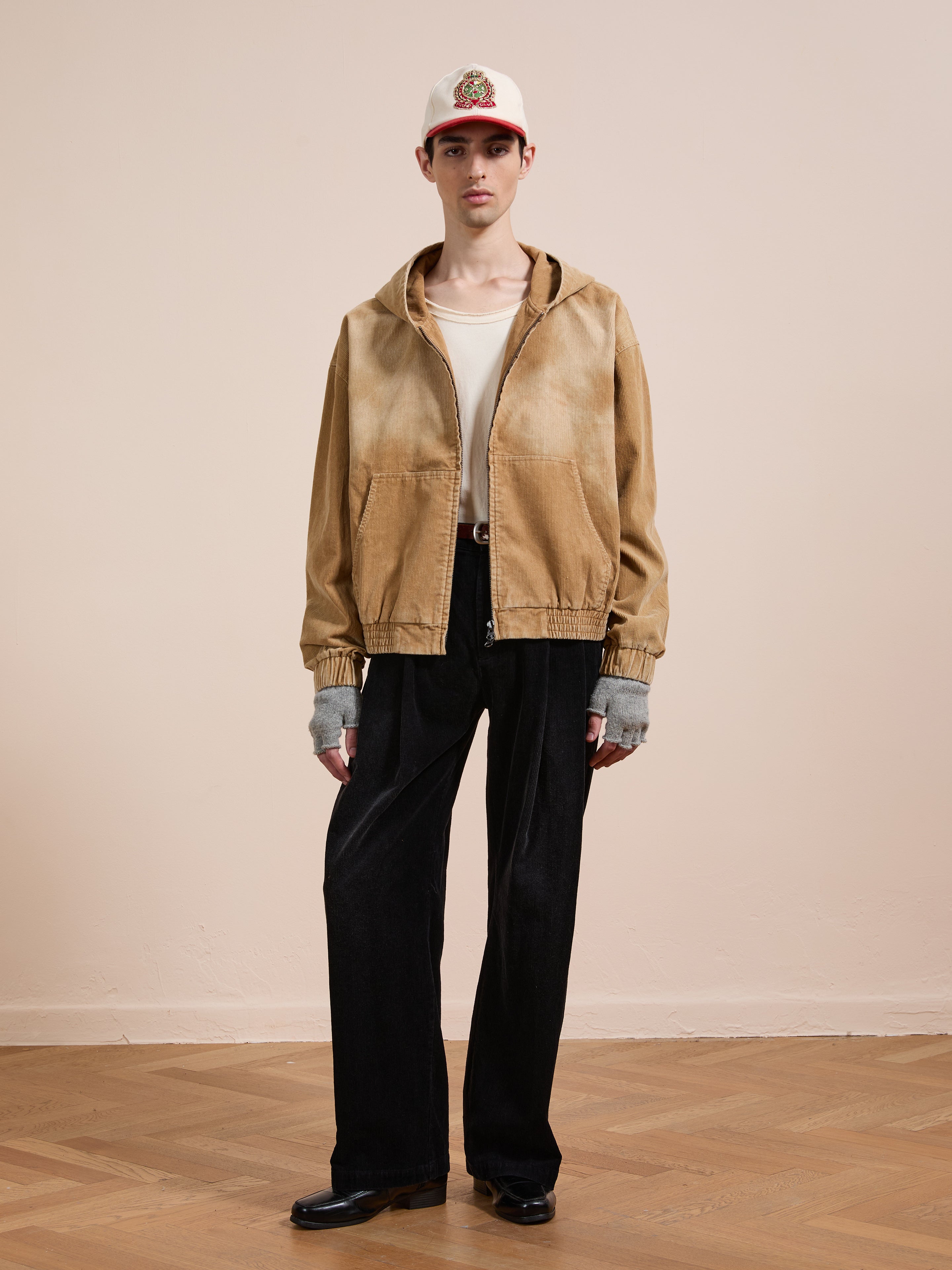 Indoors against a light wall, a person wears FOUND’s Corduroy Zip Up Hoodie in beige, black wide-leg pants and shoes, a white cap with a red brim, and fingerless gloves for a vintage look.