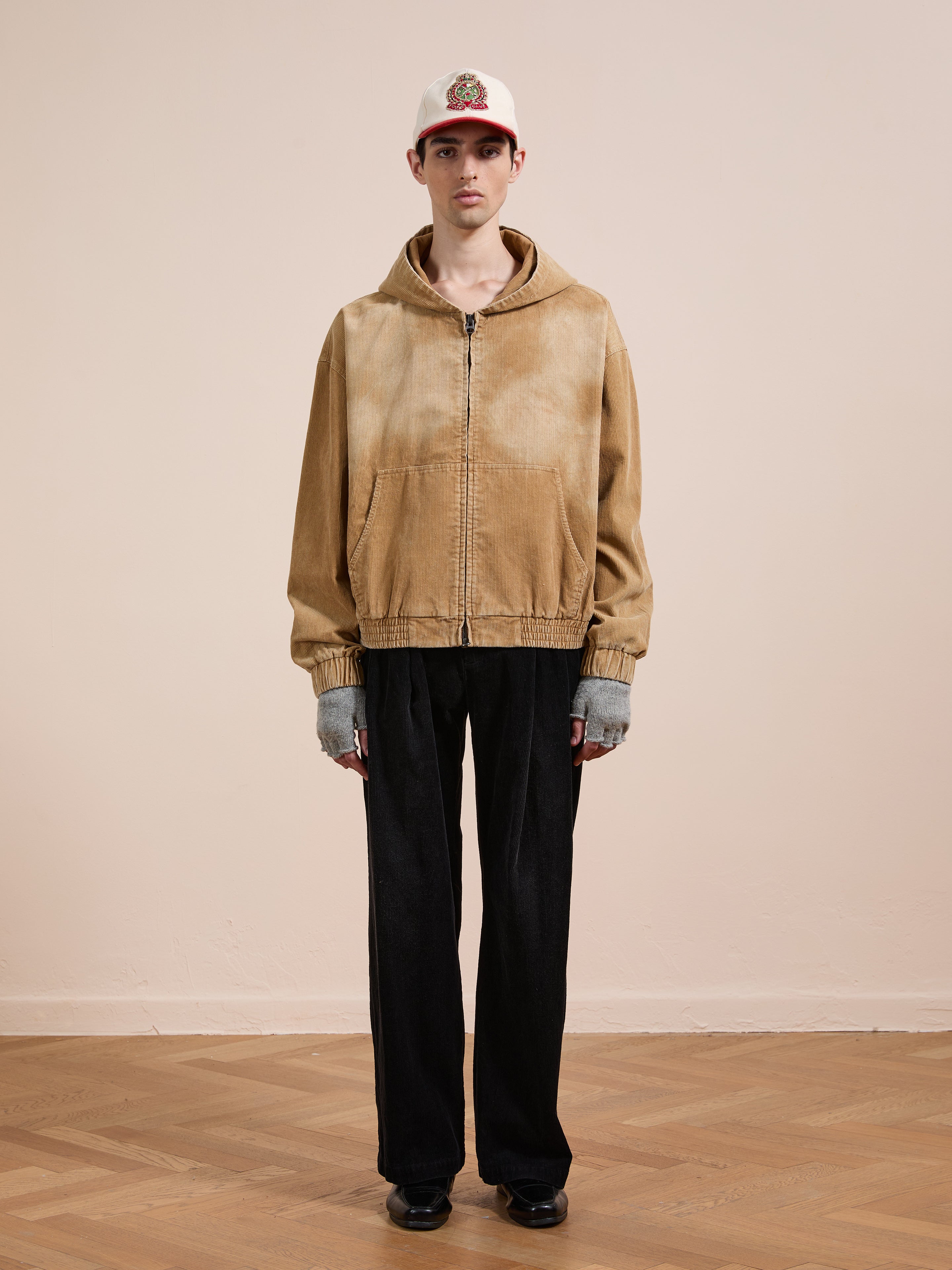 A person stands indoors against a plain background wearing the FOUND Corduroy Zip Up Hoodie in tan, styled with black pants, gray fingerless gloves, black shoes, and a white cap for a relaxed unisex look.