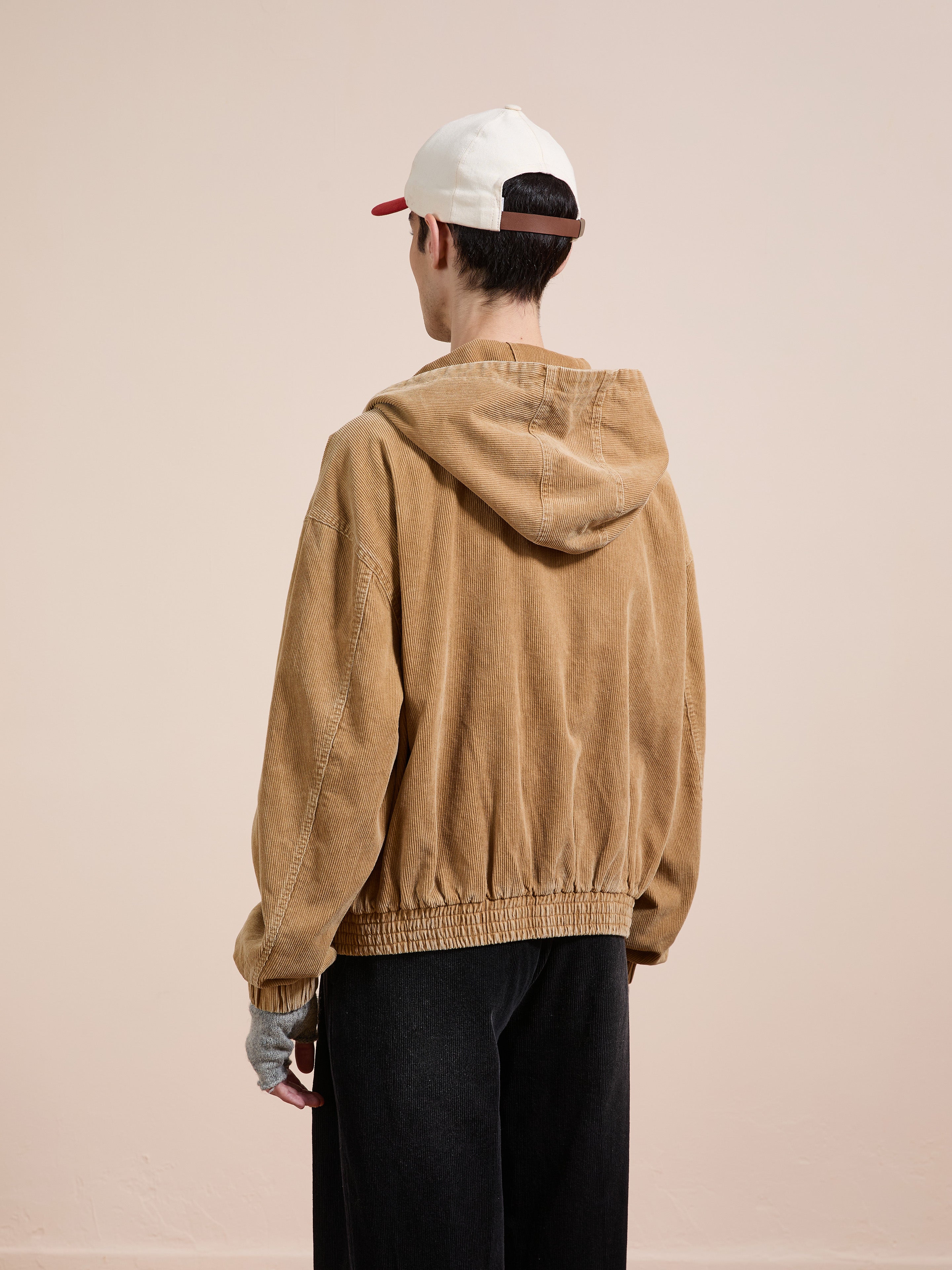 A person in a tan FOUND Corduroy Zip Up Hoodie, black pants, and a white cap with a red bill faces away from the camera against a plain light background, creating a vintage aesthetic.