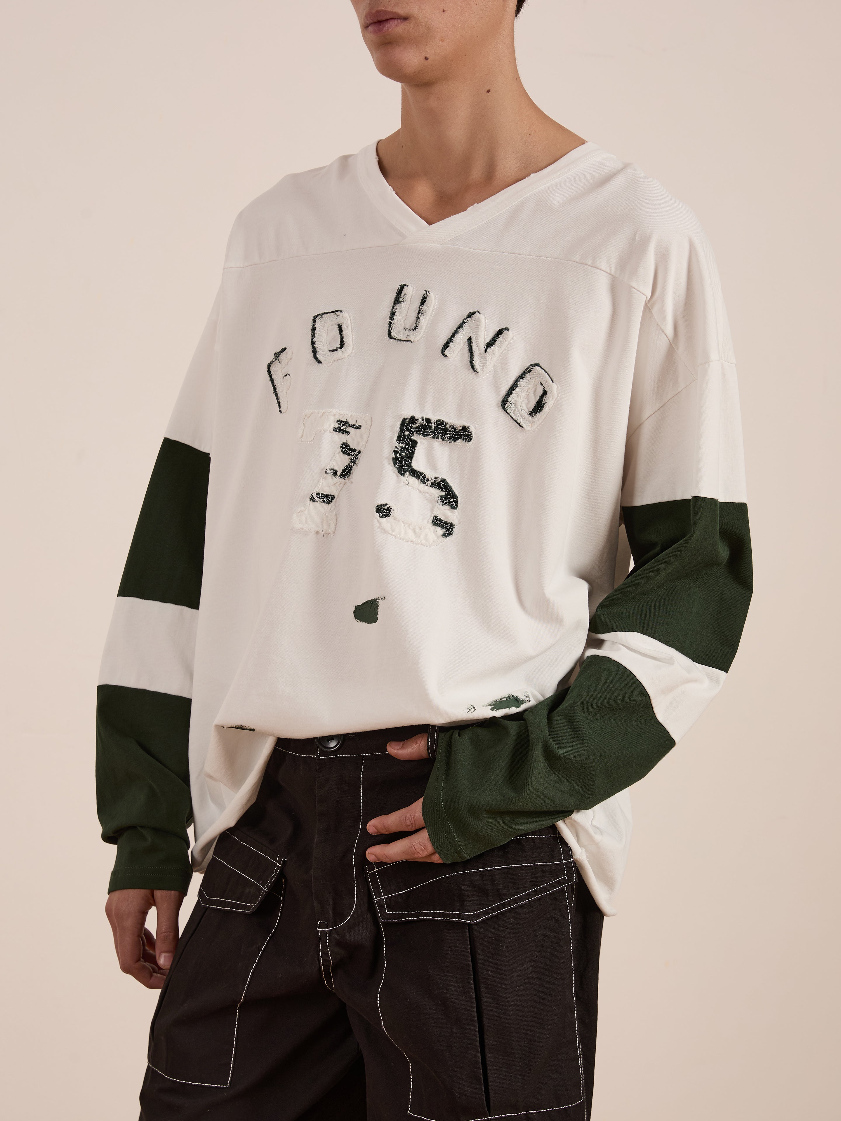 Someone wears the FOUND Distressed LS Team Jersey, a relaxed fit long-sleeve shirt in white with green hues and "FOUND 35" print, embodying vintage sportswear. It's perfectly matched with dark pants accented by white stitching.