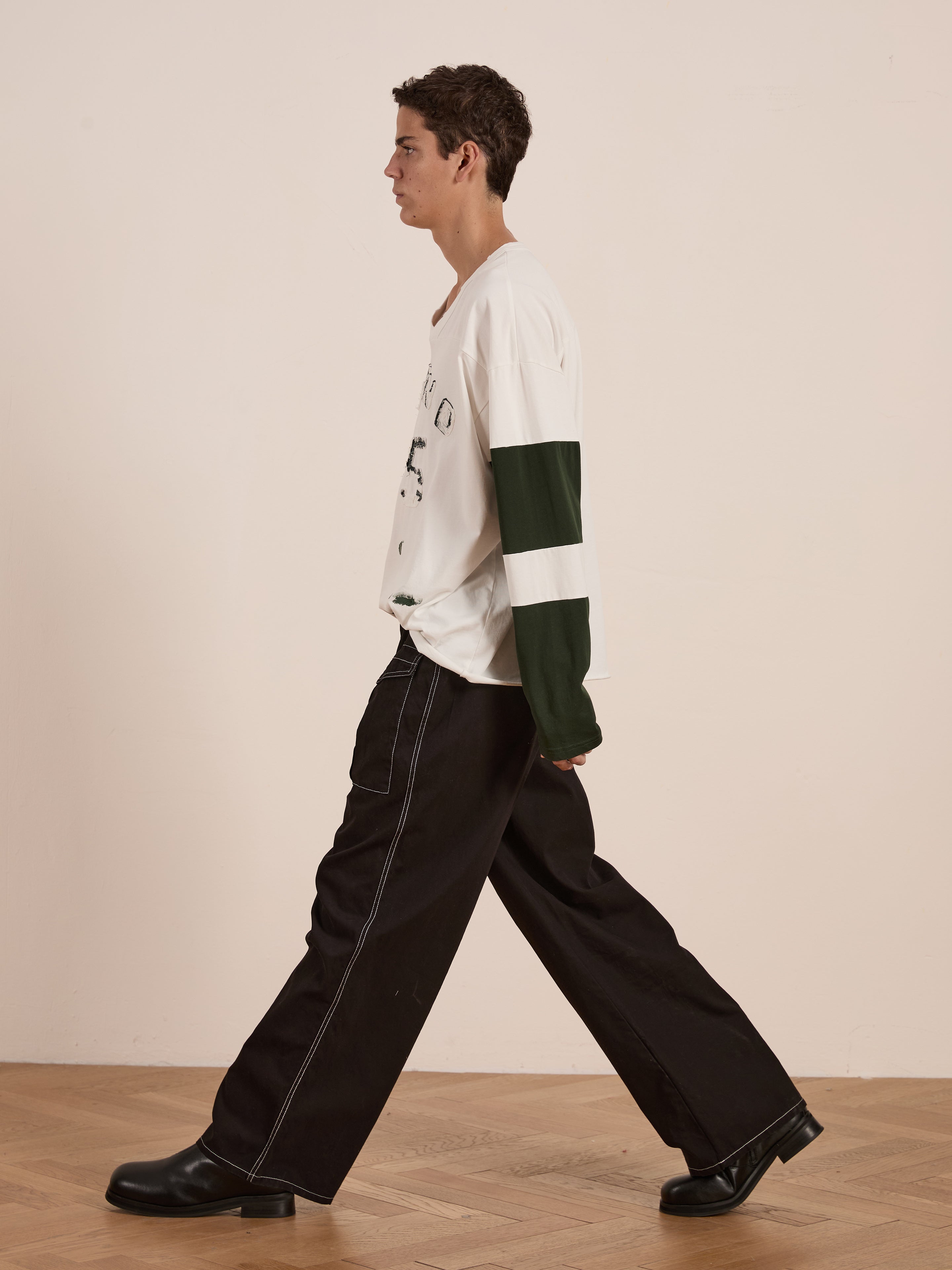 A person in profile wears the FOUND Distressed LS Team Jersey, featuring dark green sleeves, paired with black wide-leg pants with white stitching and black shoes. This outfit, set against a wooden floor, evokes a vintage sportswear vibe combining style and comfort seamlessly.