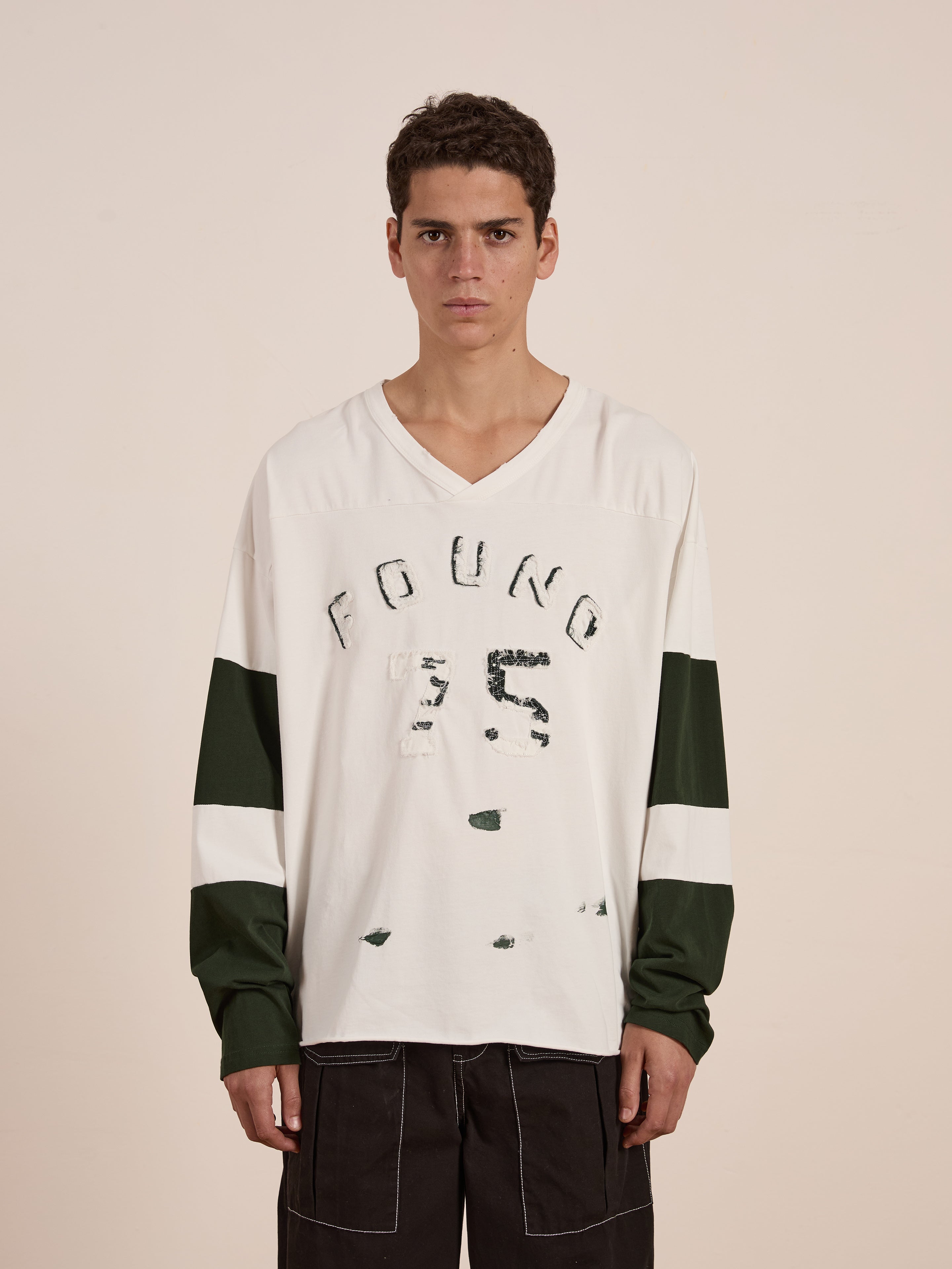 A person wears a FOUND Distressed LS Team Jersey, a relaxed-fit, distressed white and green long-sleeve shirt with "FOUND 25" on it, against a plain background.