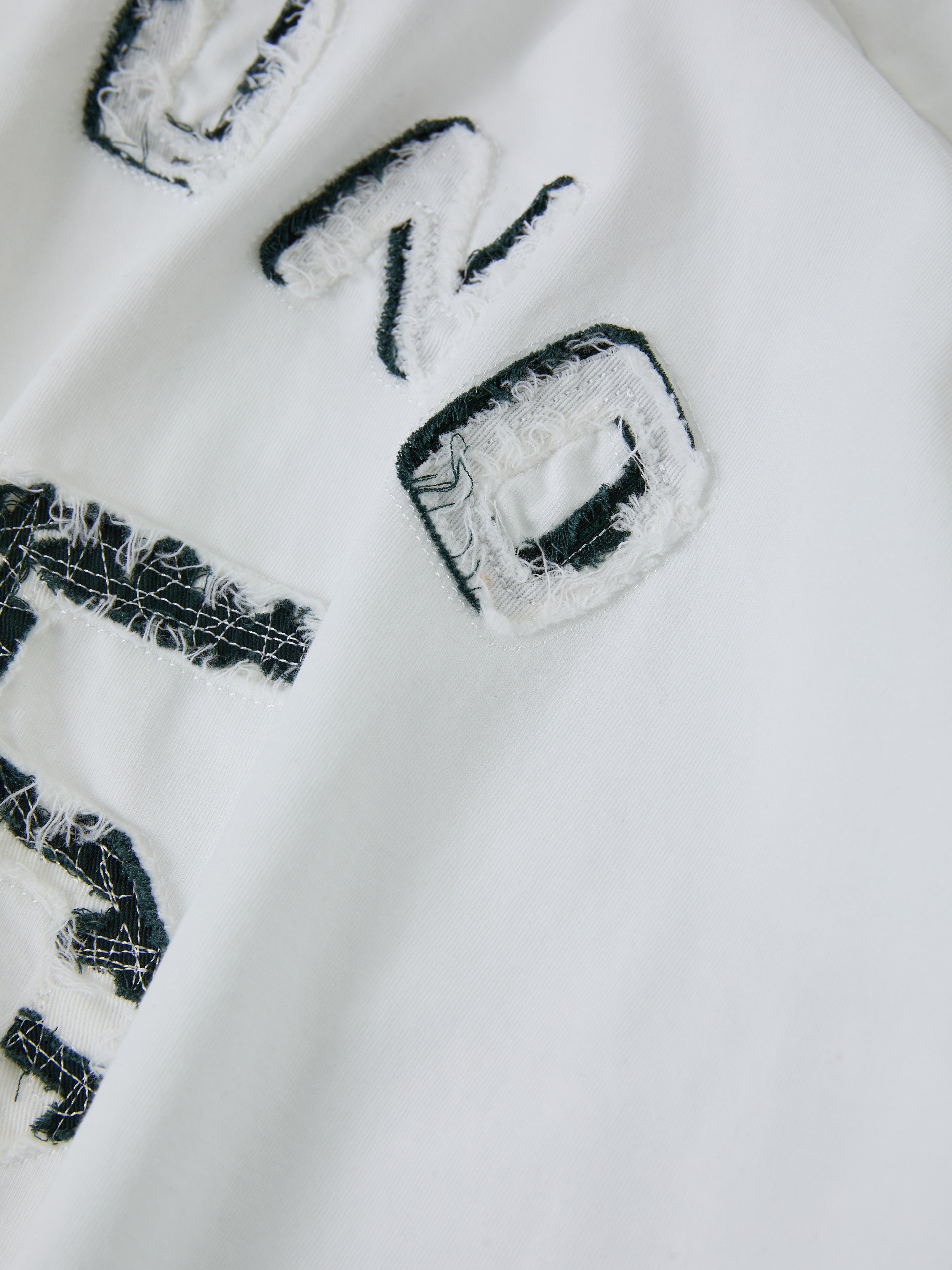 Close-up of Distressed LS Team Jersey by FOUND, showcasing its white fabric with black, frayed letters that evoke a textured, vintage sportswear look.