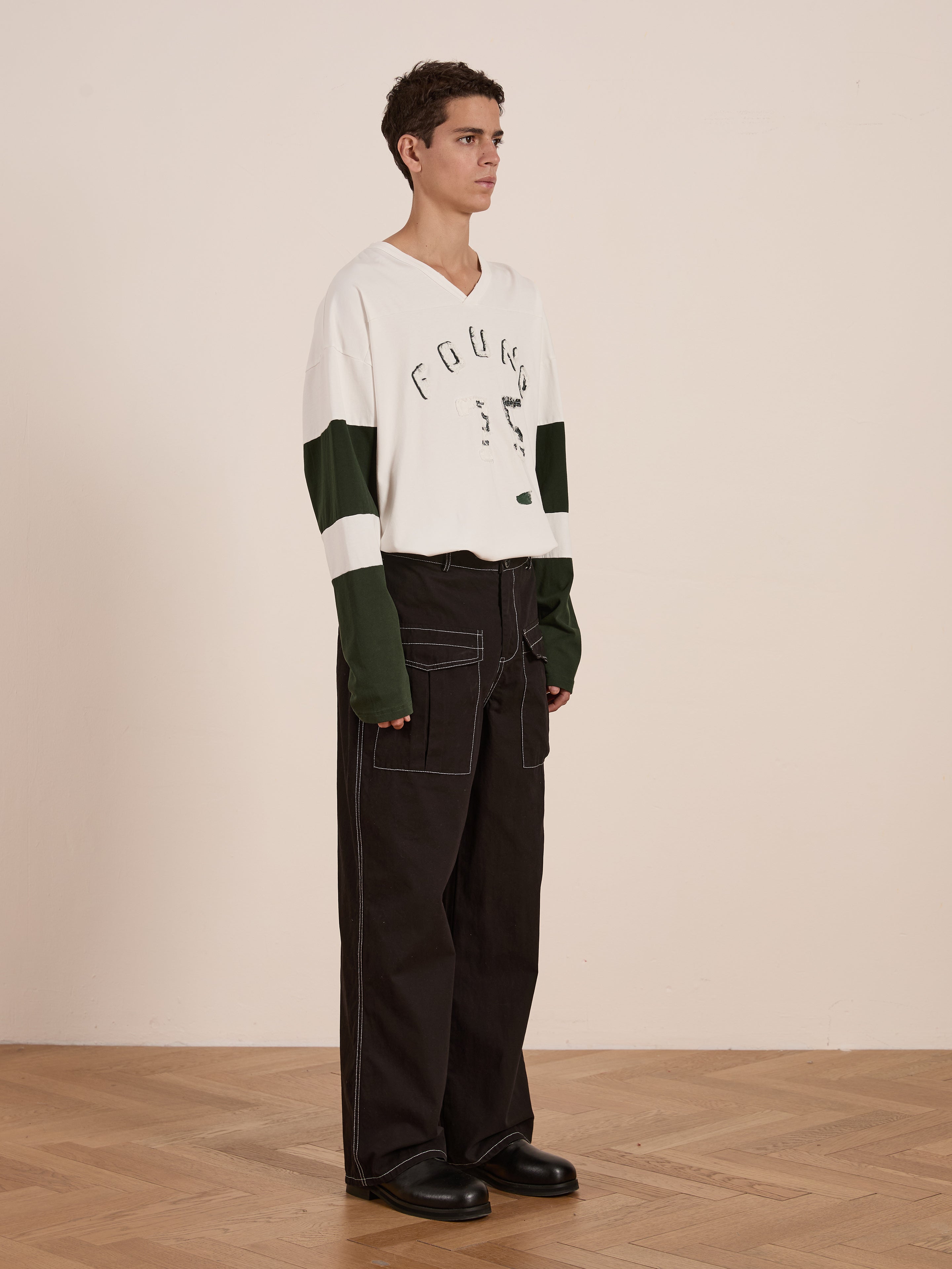 A person in a relaxed fit white and green "FOUND" Distressed LS Team Jersey, with black wide-leg pants and shoes, stands on a wooden floor against a plain wall, capturing vintage sportswear's essence.