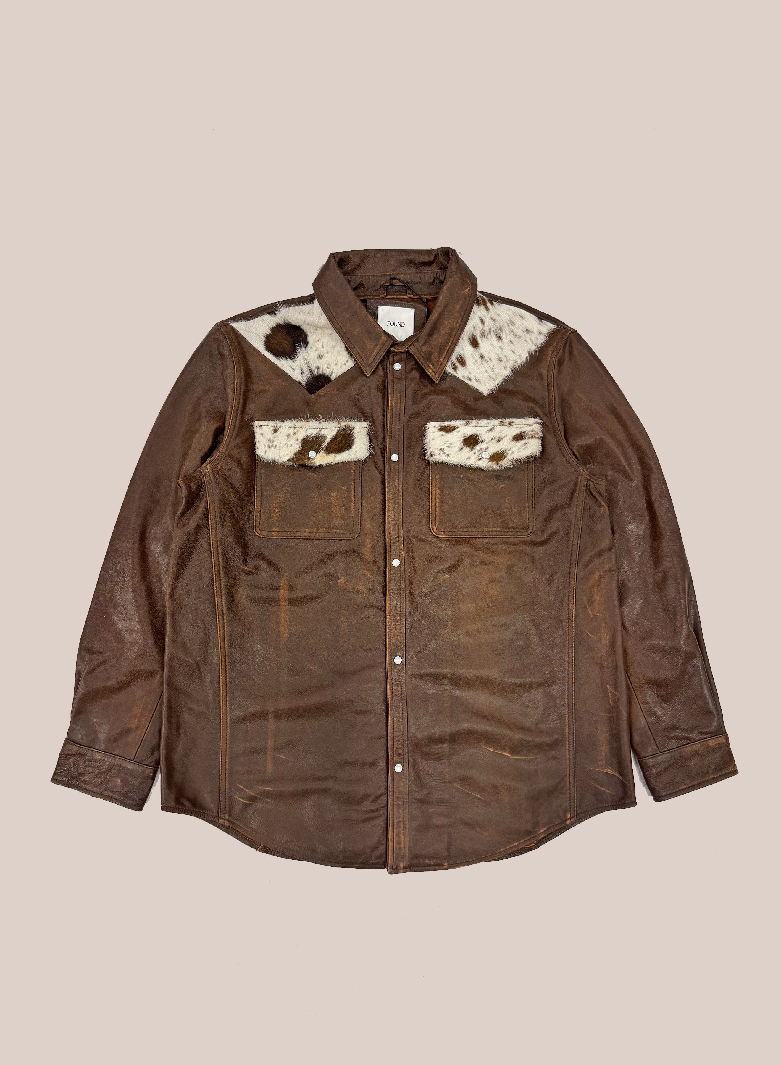 The FOUND Distressed Leather Cowhide Contrast Western Shirt features brown distressed leather, long sleeves, two front pockets, and white cowhide accents on the shoulders and upper chest. Unisex style shown laid flat on a neutral background.