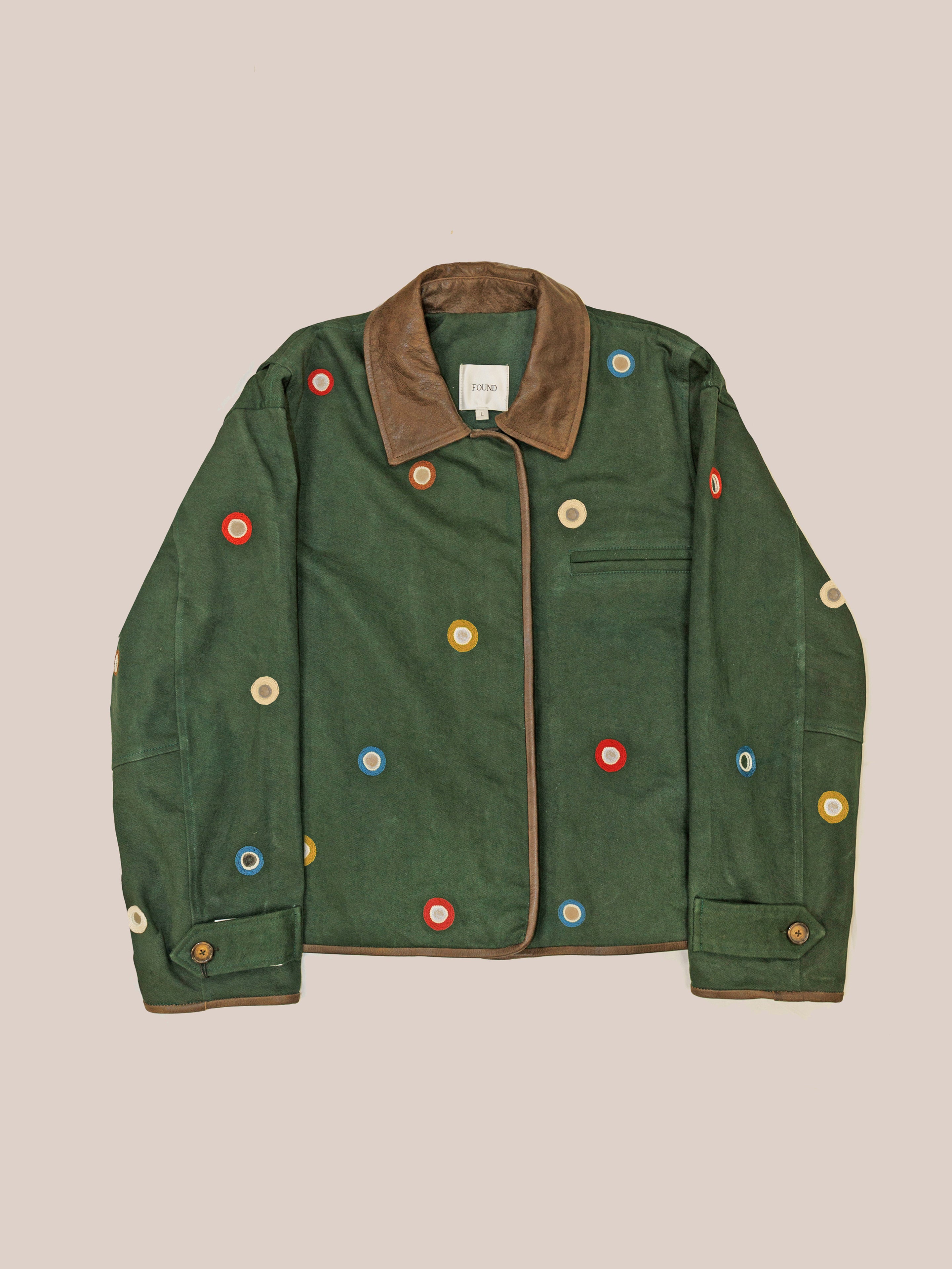 The Hand Embroidered Mirror Abhala Jacket by Found features green cotton twill, a brown collar, and colorful circular patches inspired by Sheesha Kari designs, showcased against a plain background.