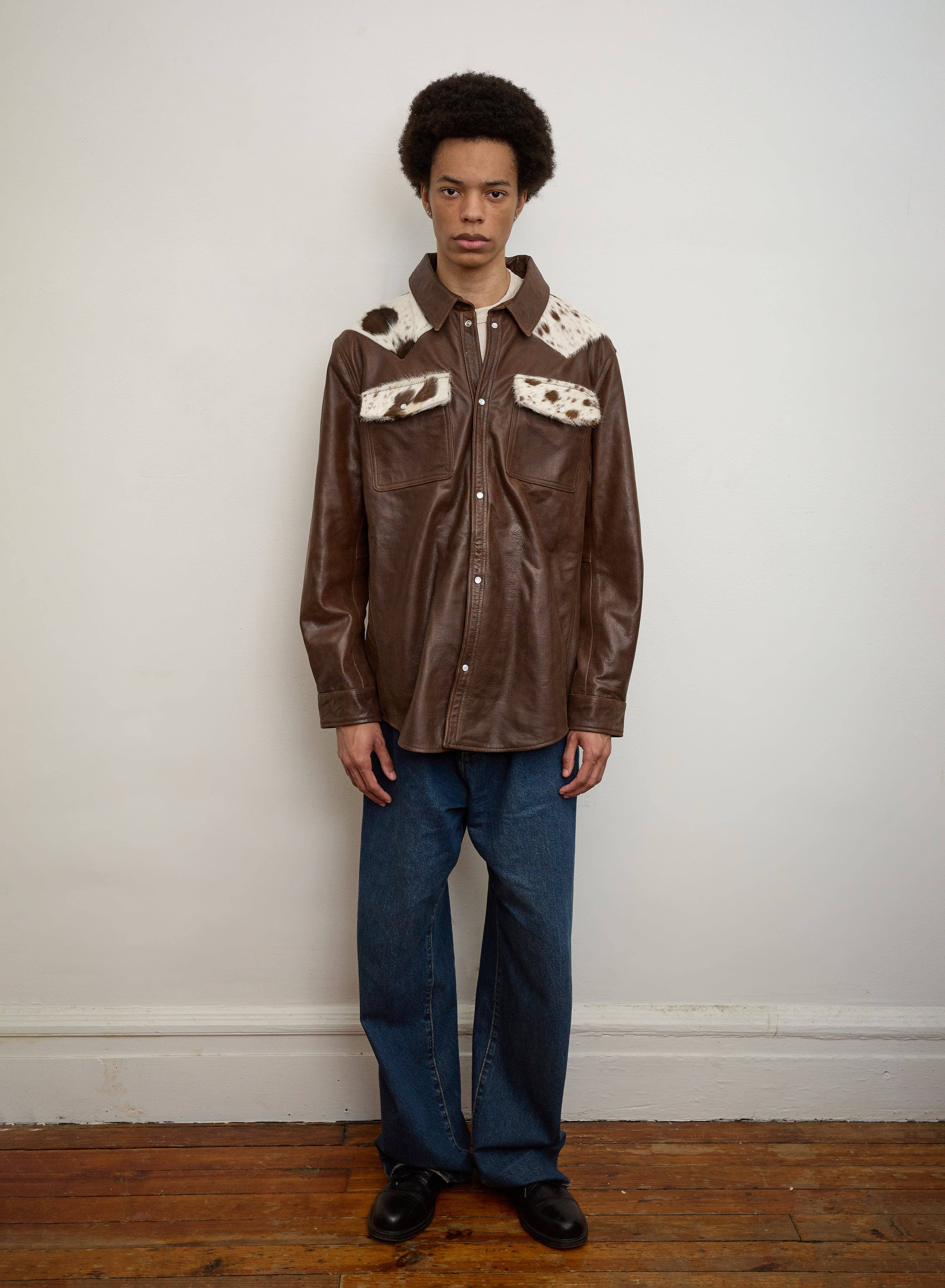 A person stands indoors against a plain wall, wearing the FOUND Distressed Leather Cowhide Contrast Western Shirt with fur accents, blue jeans, and black shoes.