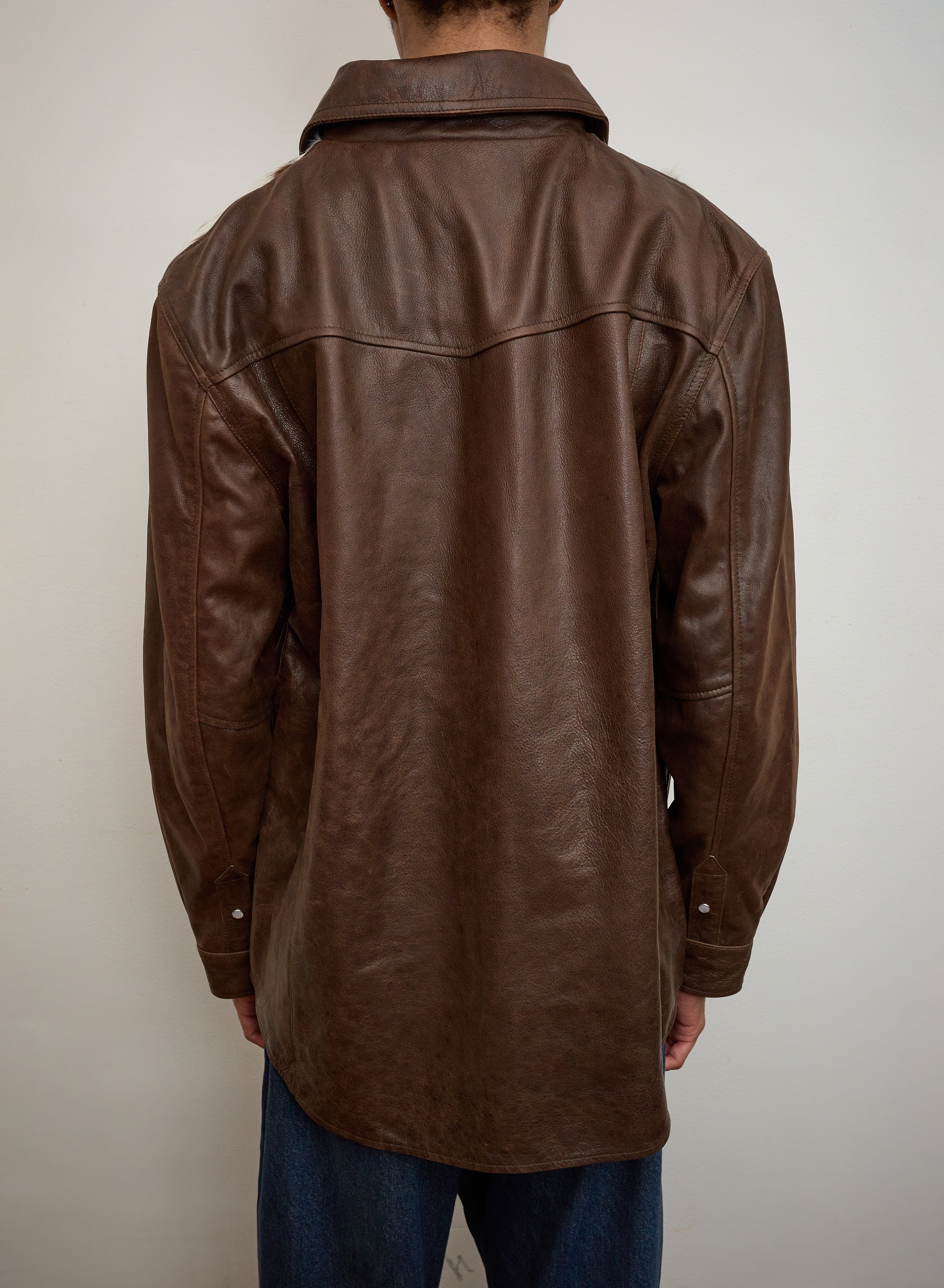 A person facing away from the camera wears a FOUND Distressed Leather Cowhide Contrast Western Shirt and blue jeans against a plain, light-colored background.