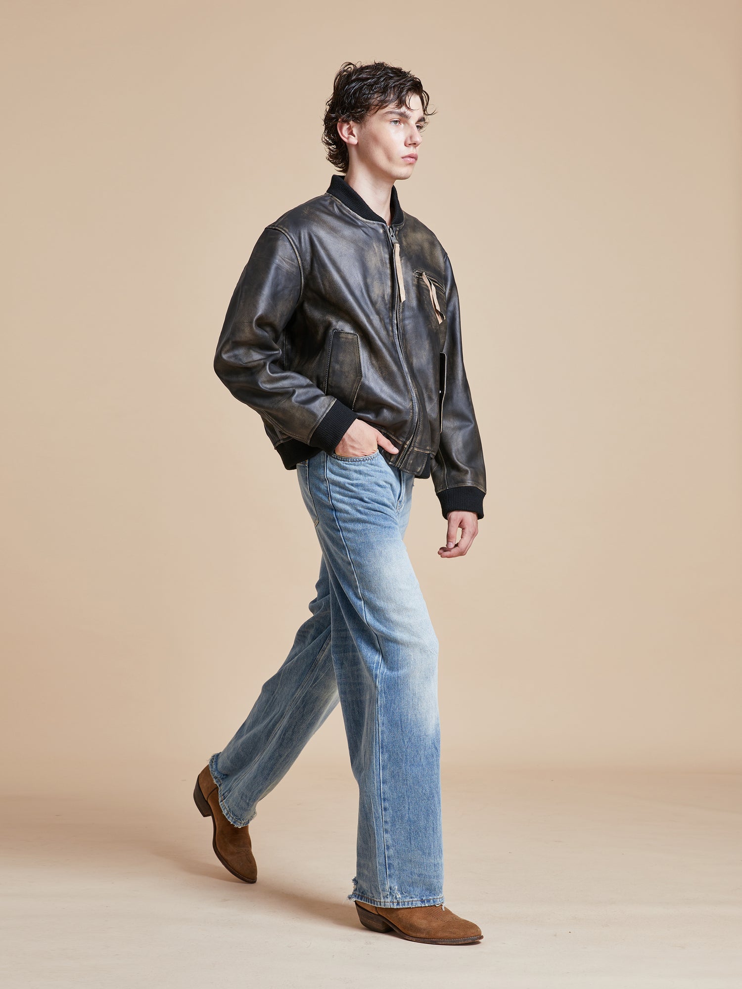A man in a Distressed Pavement Leather Bomber Jacket by Found and jeans walking on a beige background.