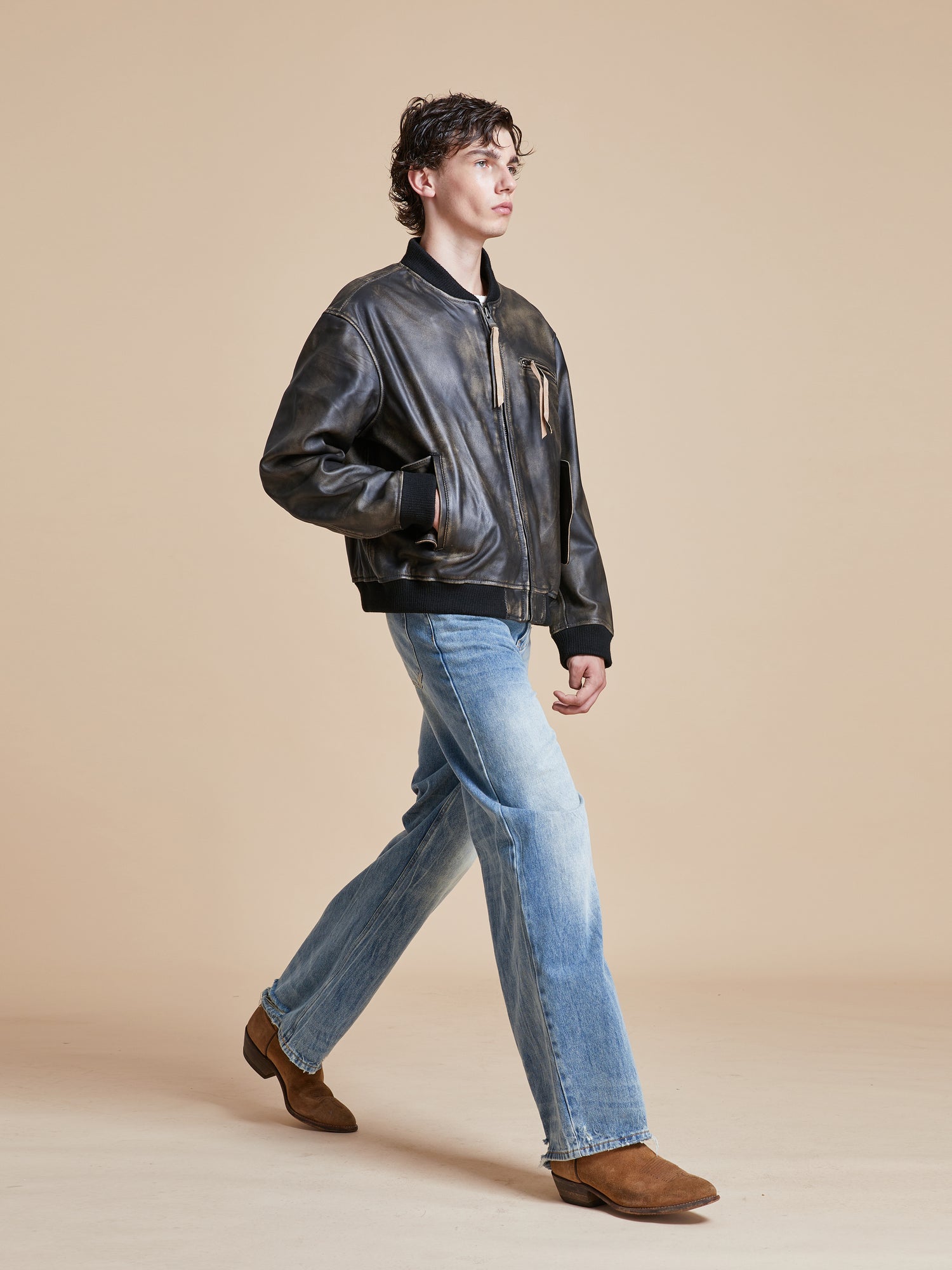 A man in jeans and a Found Distressed Pavement Leather Bomber Jacket walking on a beige background.