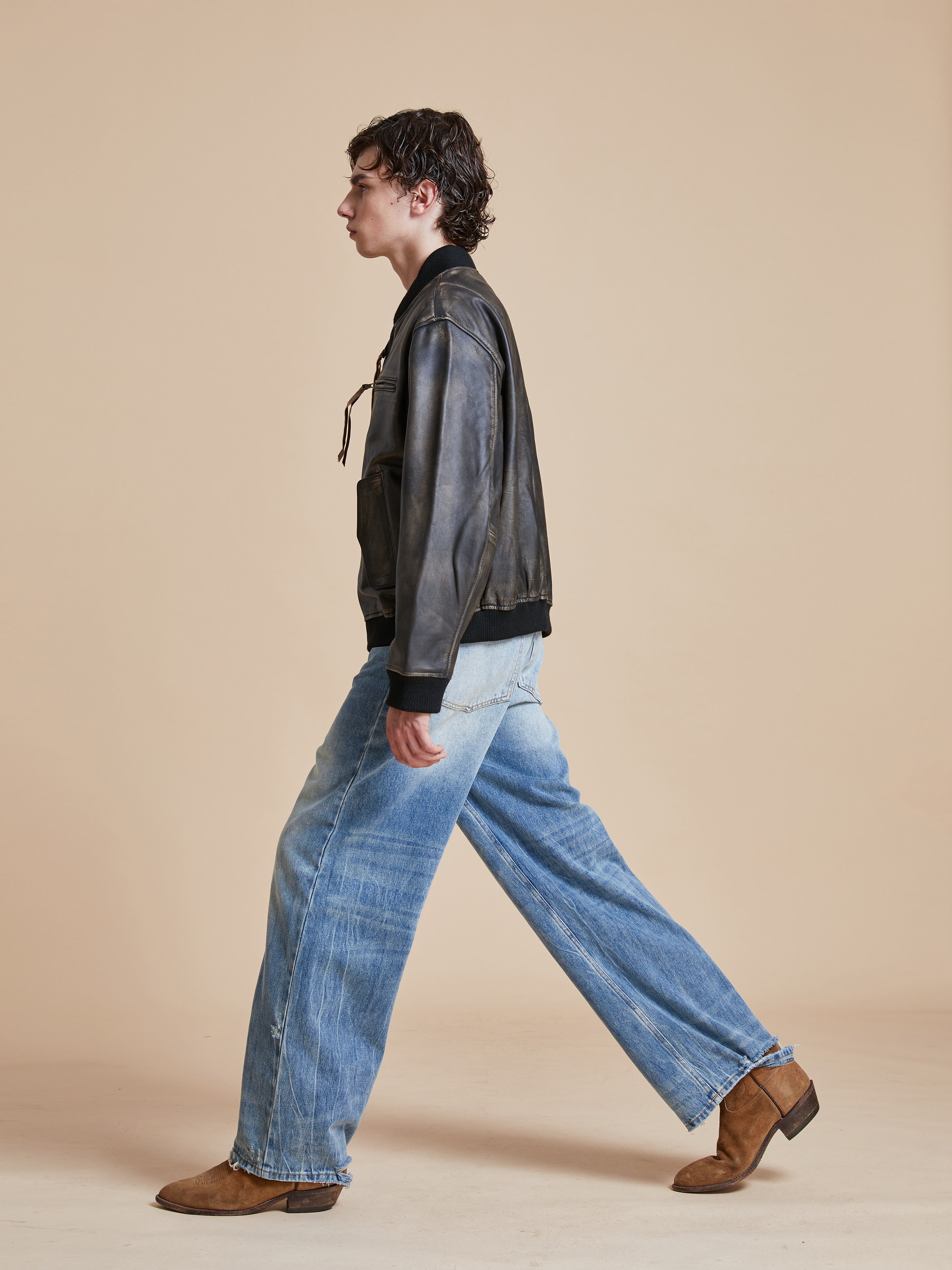 A man in jeans and a Found Distressed Pavement Leather Bomber Jacket walking on a beige background.