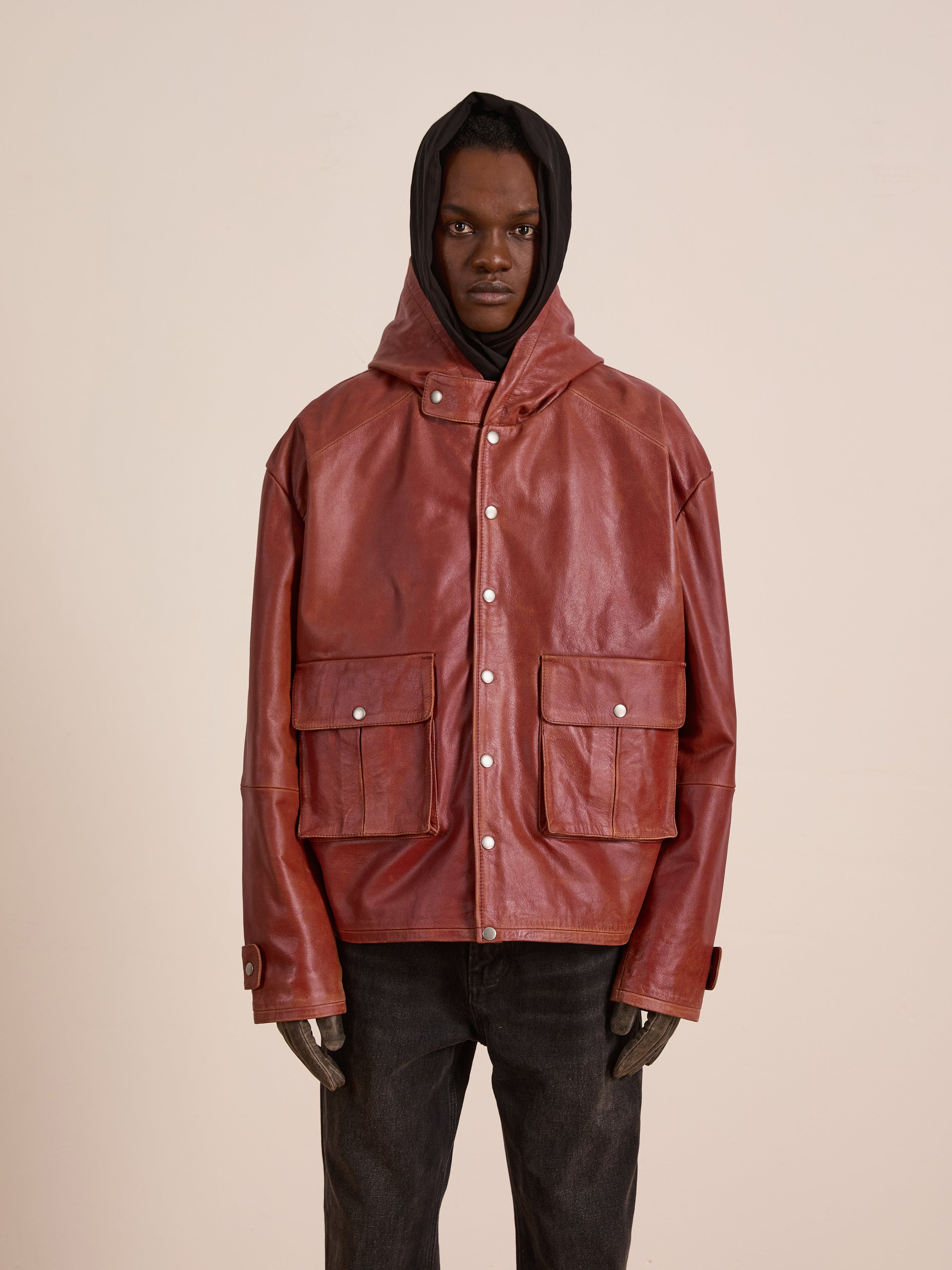 A person models the FOUND Distressed Leather Pocket Hoodie—vintage red genuine cowhide with front pockets and black hood—paired with dark jeans and gloves, standing against a plain beige background.