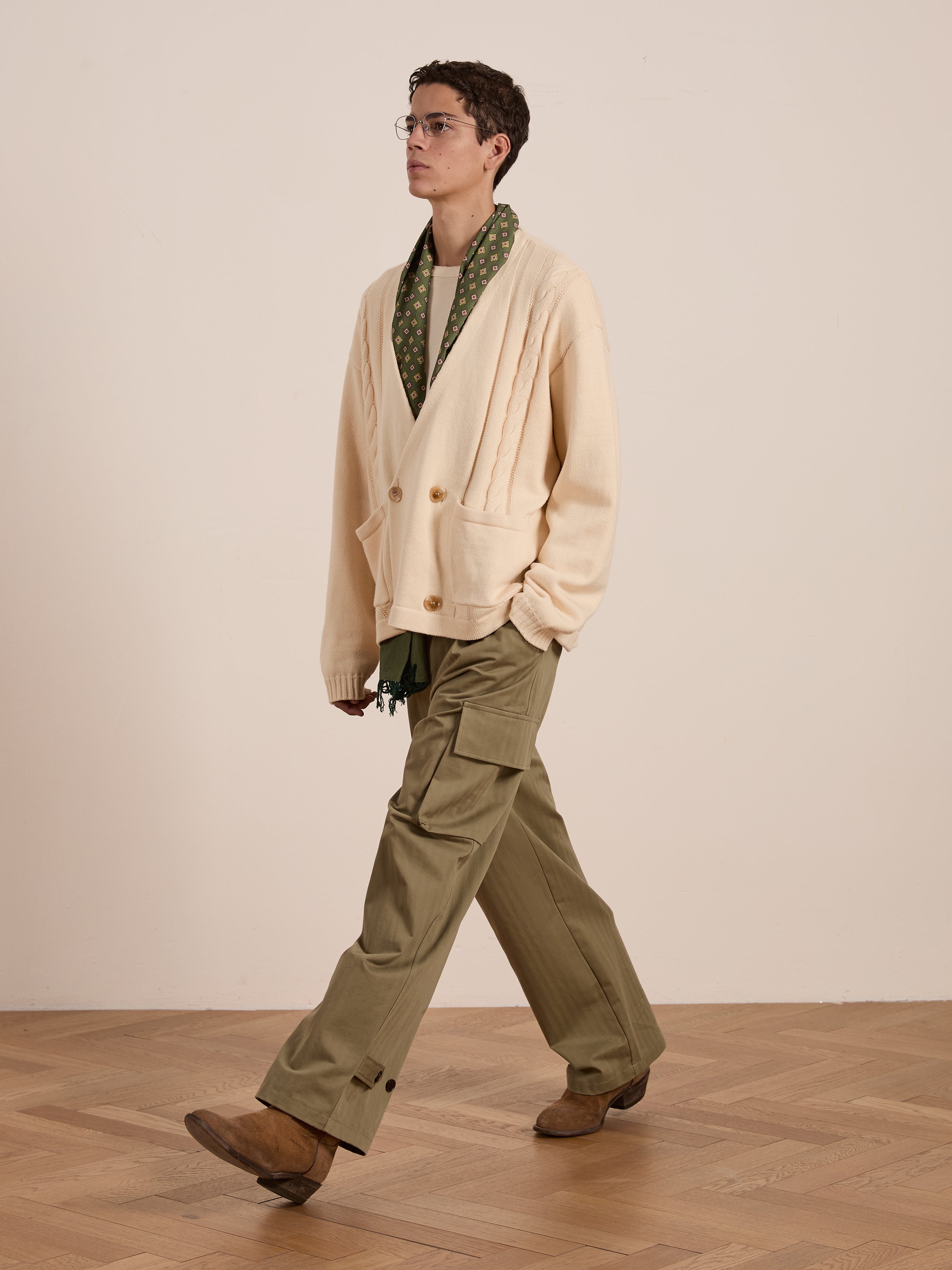 A person in glasses walks across a wooden floor, sporting a unisex cream Double Breasted Knit Cardigan by FOUND, paired with a green scarf, khaki cargo pants, and brown shoes.