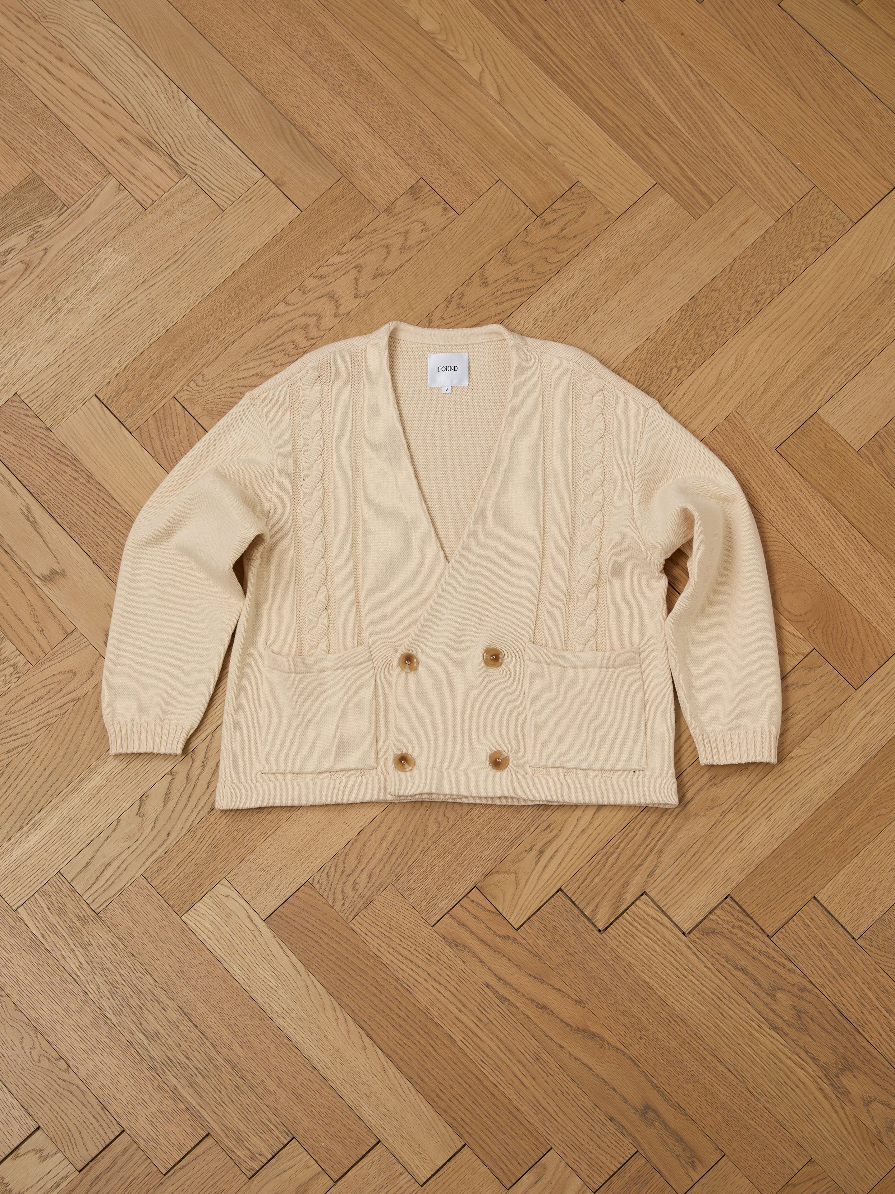 A FOUND Double Breasted Knit Cardigan in beige, made from a cozy cotton blend with two front pockets, is displayed flat on a herringbone wooden floor.