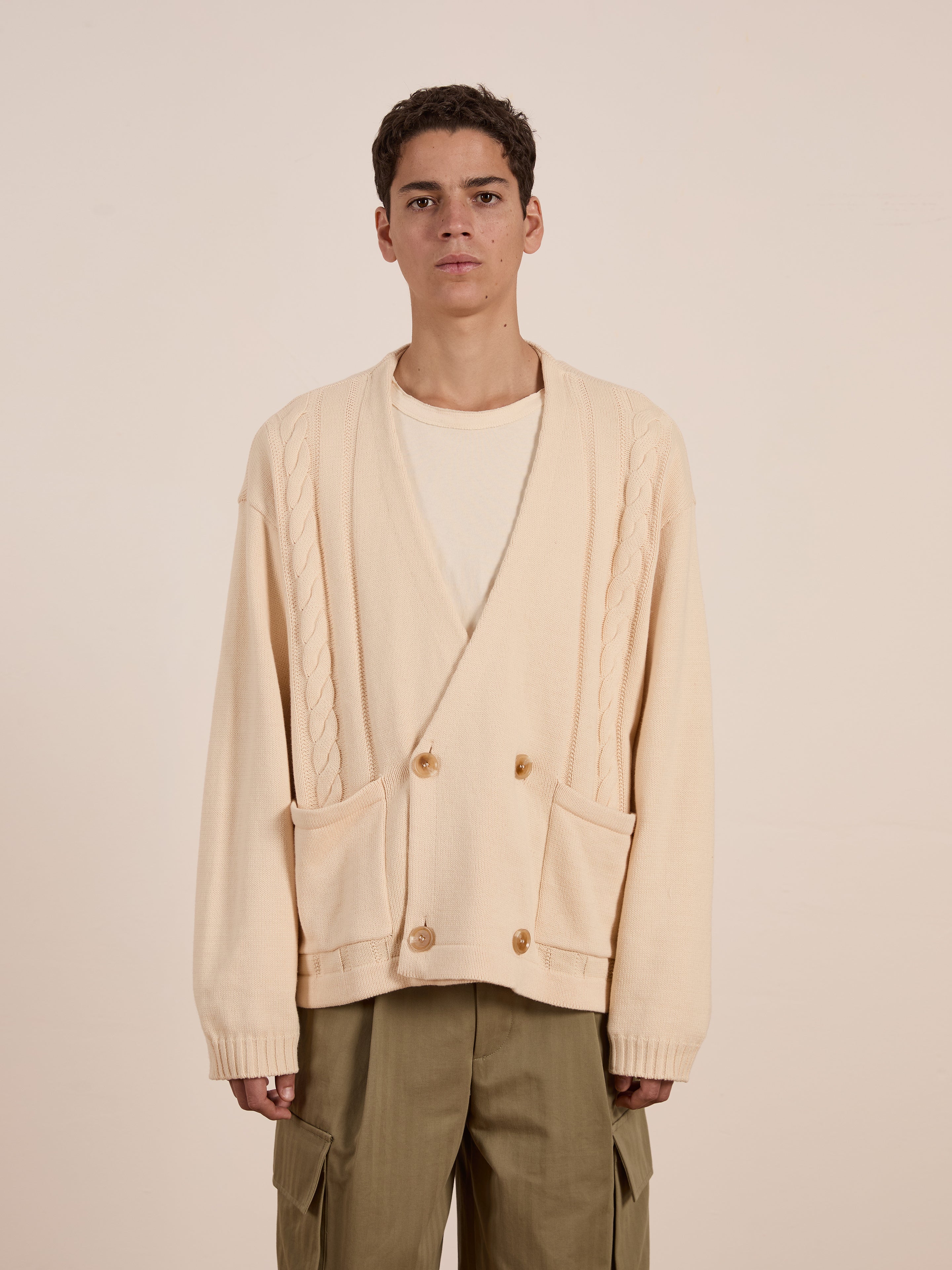 A person wearing a cream-colored Double Breasted Knit Cardigan from FOUND—a relaxed-fit, midweight cotton blend with buttons and pockets—over a white shirt and olive cargo pants, against a plain background.