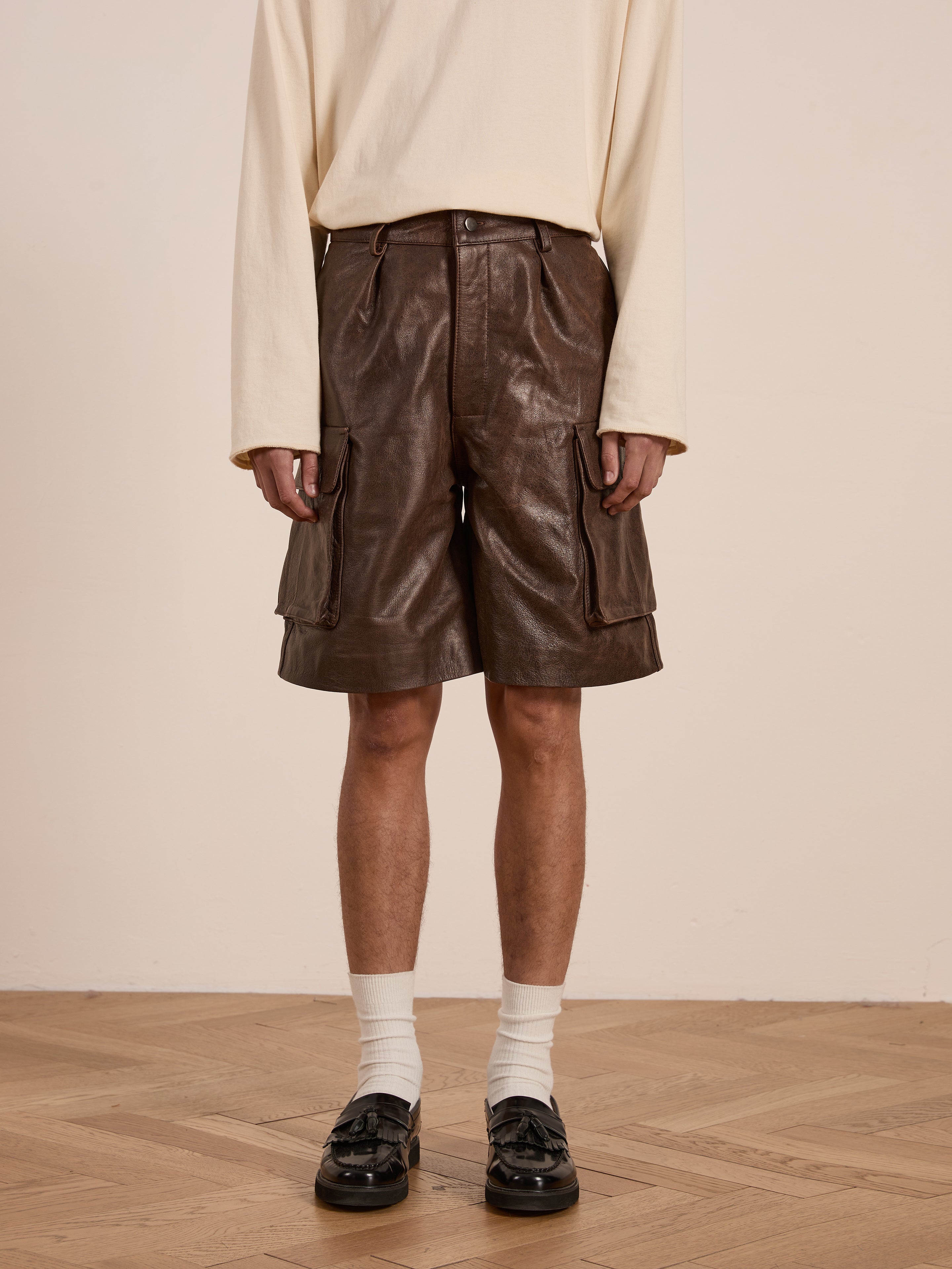 On a wooden floor, someone in a cream long-sleeve shirt and Dusk Distressed Genuine Leather Cargo Shorts by Found stands wearing black loafers and white socks, highlighting an unisex relaxed fit crafted from ethically sourced materials.