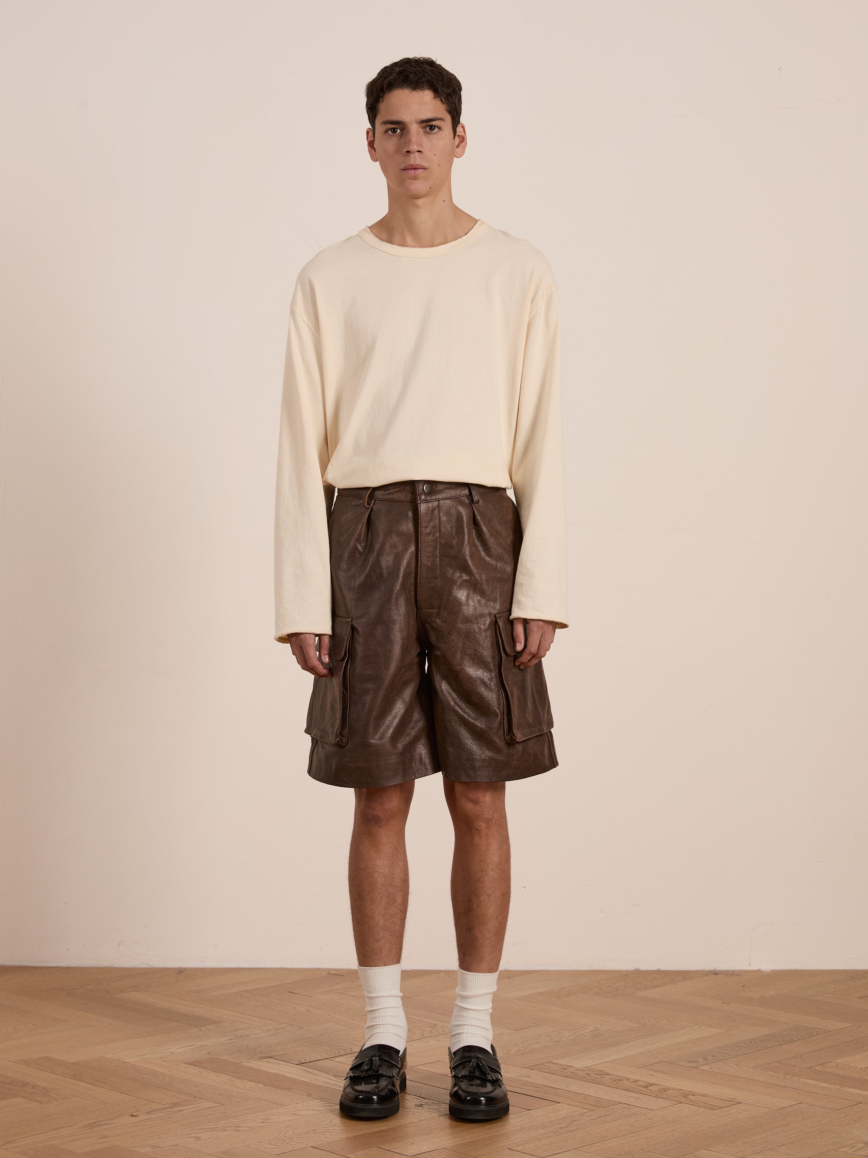 A person is against a plain backdrop wearing a cream long-sleeve shirt, Dusk Distressed Genuine Leather Cargo Shorts by Found, white socks, and black shoes. The unisex relaxed fit ensemble features ethically sourced materials for an eco-conscious style statement.