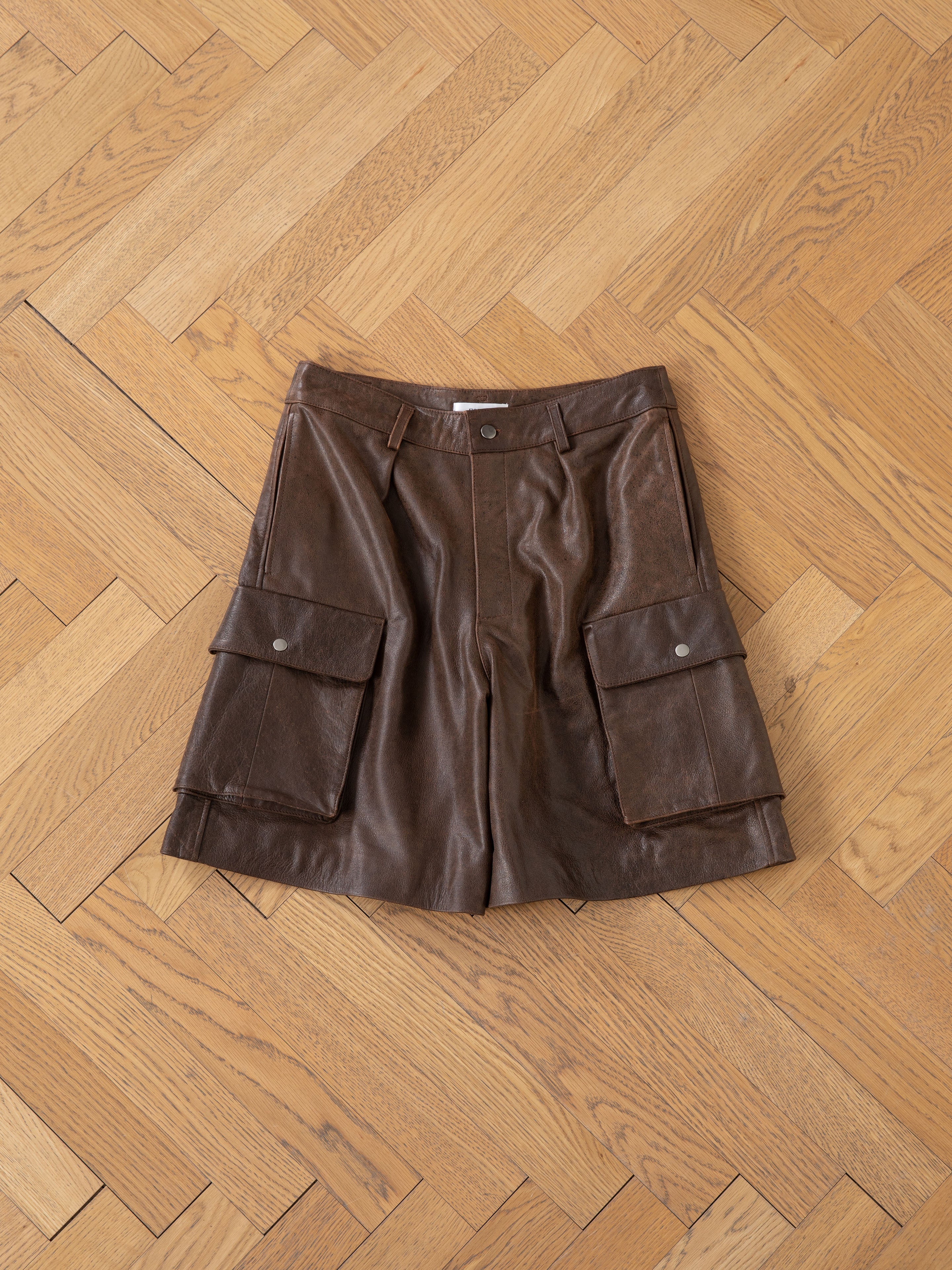 The Dusk Distressed Genuine Leather Cargo Shorts by Found, made from ethically sourced materials, feature large front pockets and are laid flat on a wooden herringbone floor.