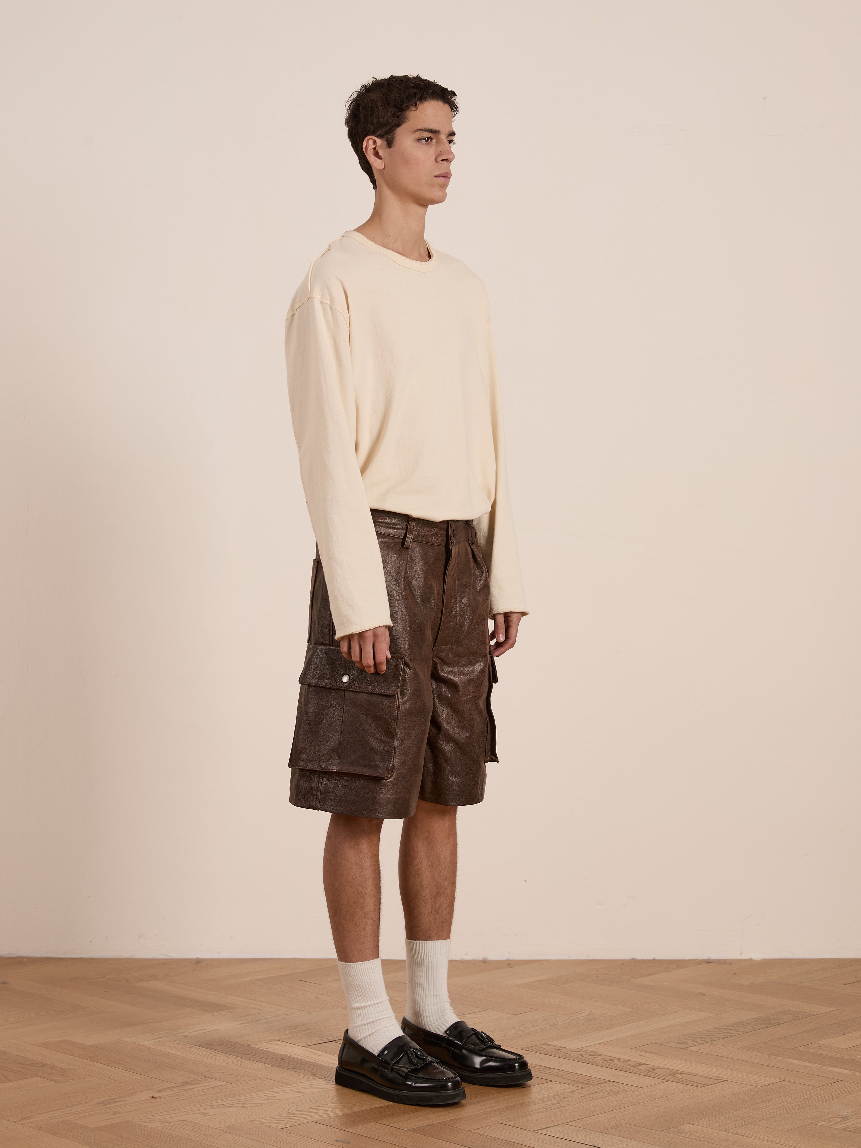 A young man in a beige long-sleeve shirt and Found's Dusk Distressed Genuine Leather Cargo Shorts stands on a wooden floor against a plain wall. His unisex, relaxed fit is completed with white socks and black loafers, all made from ethically sourced materials for a conscious style statement.