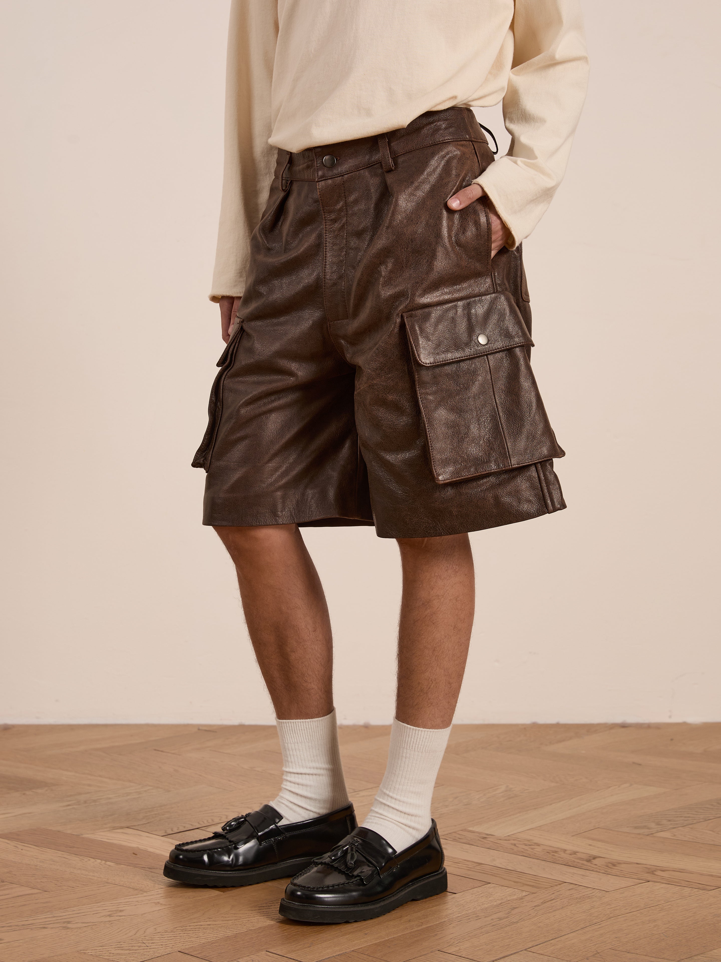 A person stands on a wooden floor, wearing a beige top and Found's Dusk Distressed Genuine Leather Cargo Shorts with side pockets. The look is completed with white socks and shiny black loafers, made from ethically sourced materials for an unisex relaxed fit.