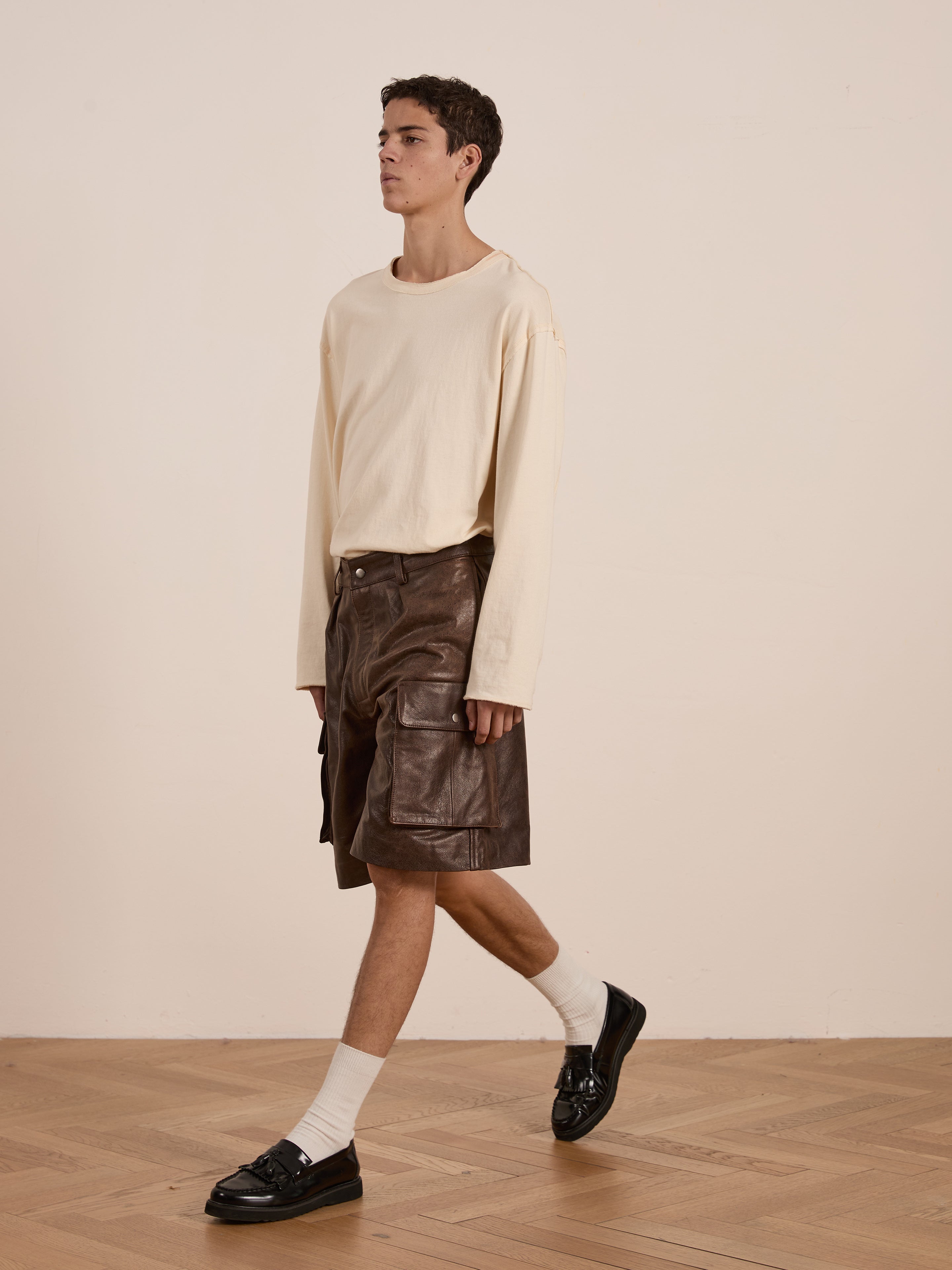 A person walks indoors in a beige long-sleeve shirt and Found's Dusk Distressed Genuine Leather Cargo Shorts, featuring a unisex relaxed fit, complemented by white socks and black loafers. The wood floor and plain wall enhance the look crafted from ethically sourced materials.