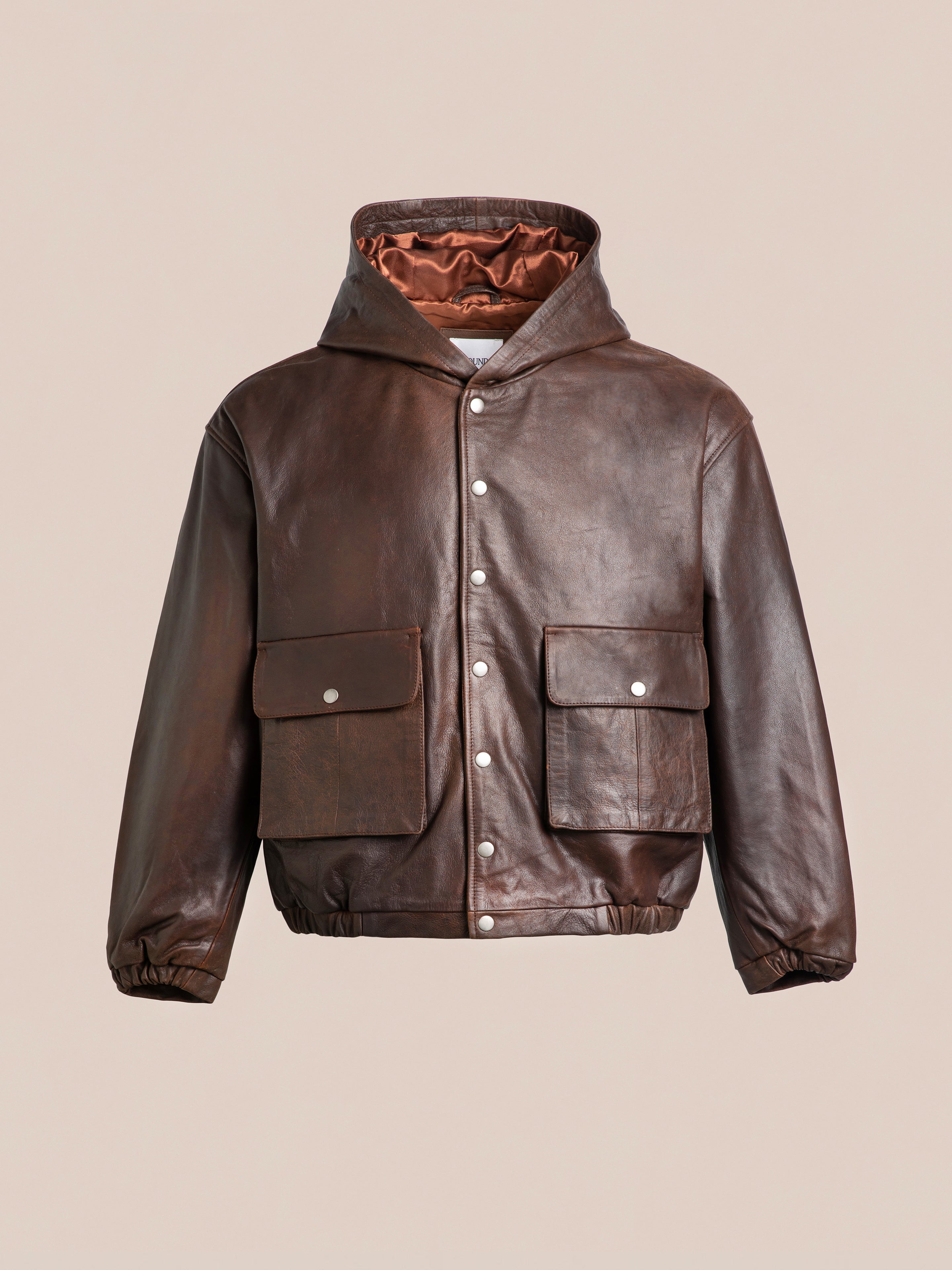 The Dusk Distressed Genuine Leather Hoodie by FOUND boasts a brown tone with a hood, front snap buttons, and two large flap pockets; this unisex jacket is finished with a gathered hem and cuffs for added style.