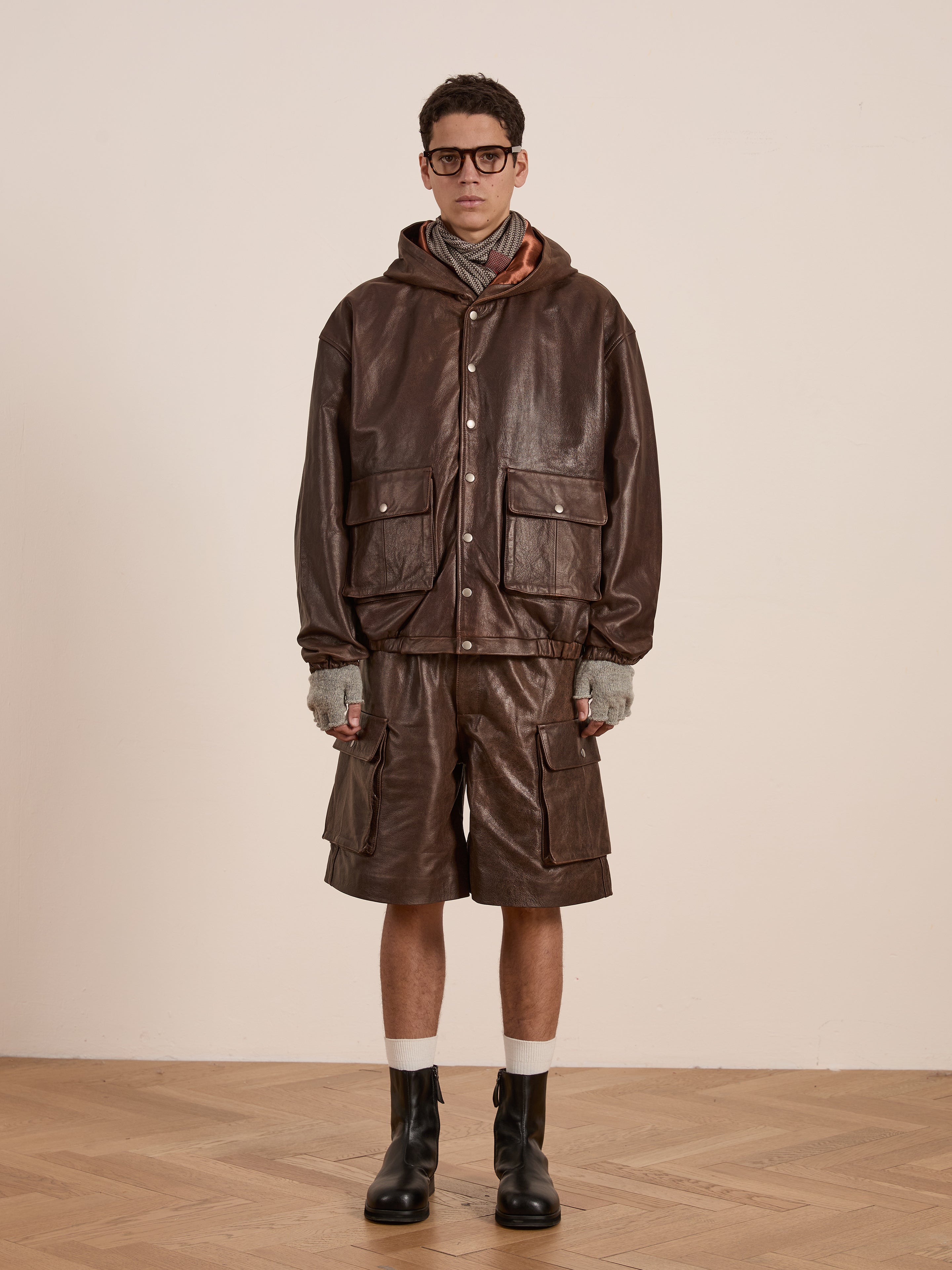 A person in FOUND's Dusk Distressed Genuine Leather Hoodie, shorts, and boots stands on a wooden floor against a simple backdrop, highlighting an outfit made from ethically sourced materials.