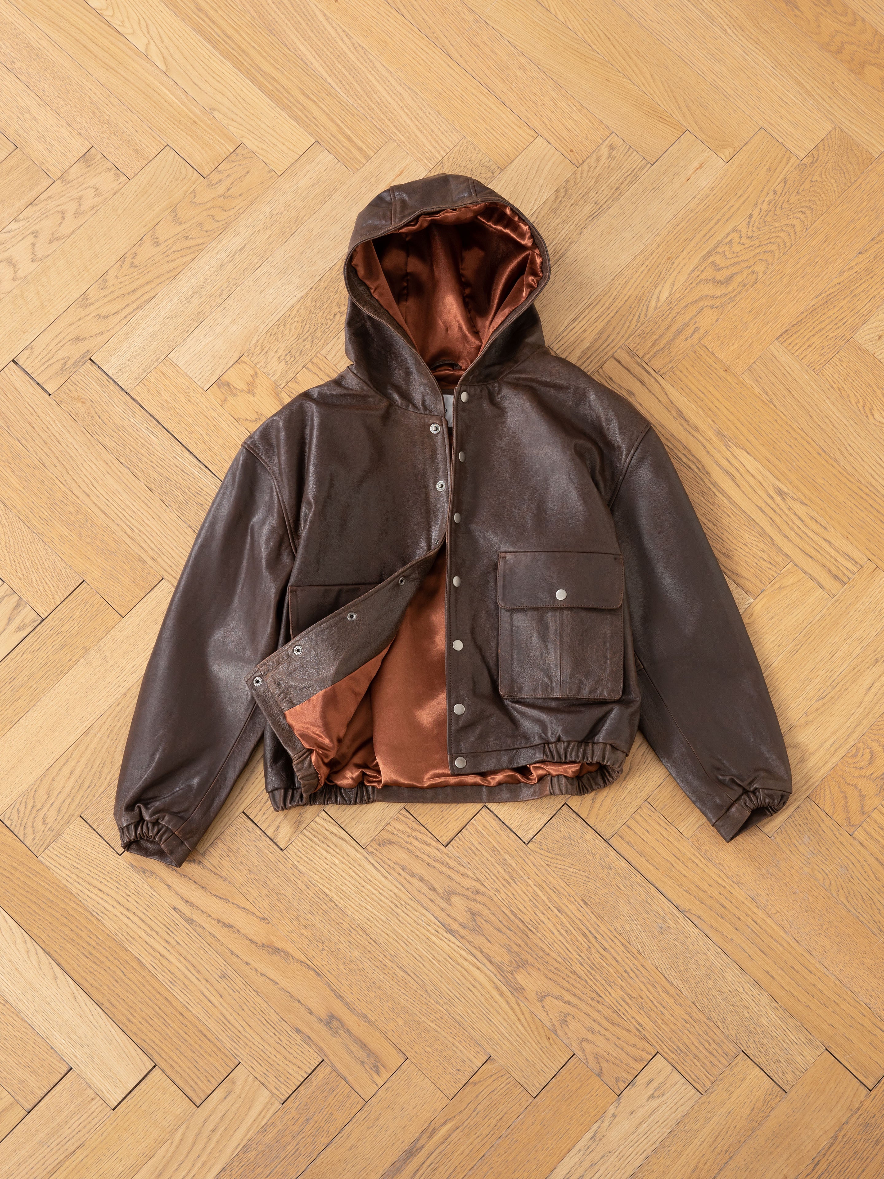 The Dusk Distressed Genuine Leather Hoodie by FOUND, in rich brown with a vibrant orange lining and hood, is elegantly displayed on a herringbone wooden floor.